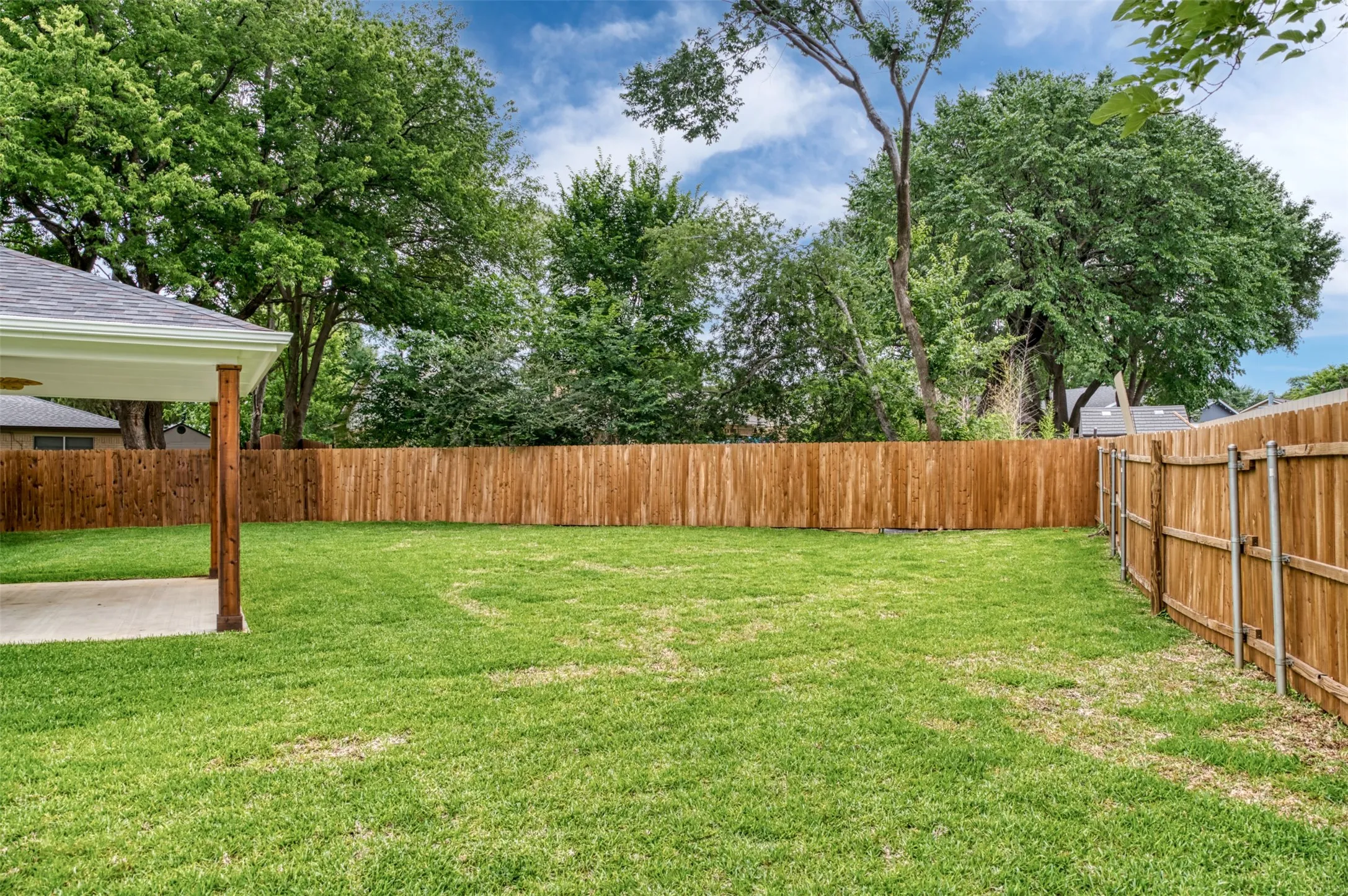 Expansive lawn area with a wood privacy fence, concrete patio, and a covered porch with wood support beams