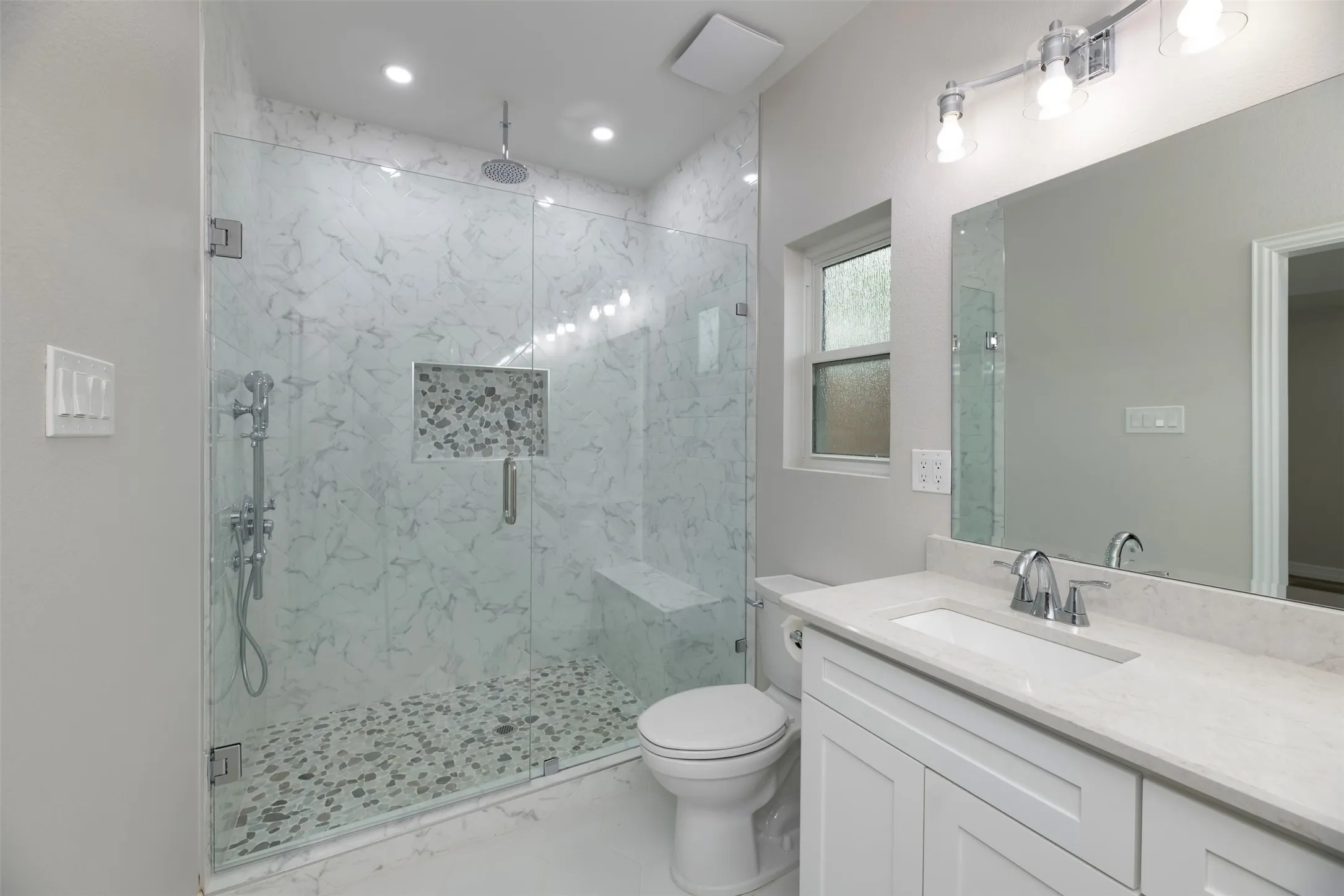 Glass-enclosed shower with a rainfall showerhead, tiled bench, and pebble-finish floor
