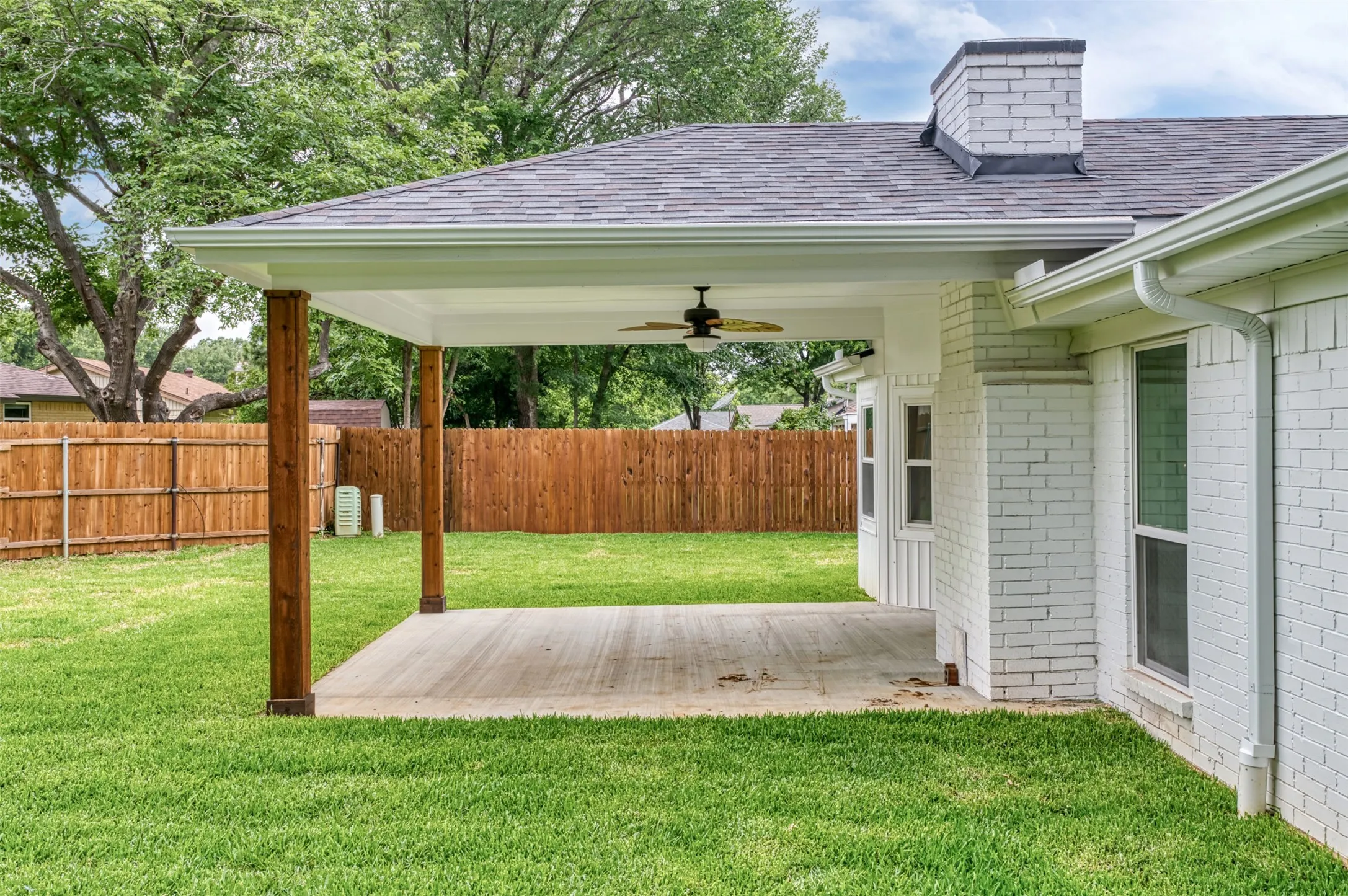Expansive backyard featuring a covered patio, wood-finish support columns, white brick exterior, and a privacy fence