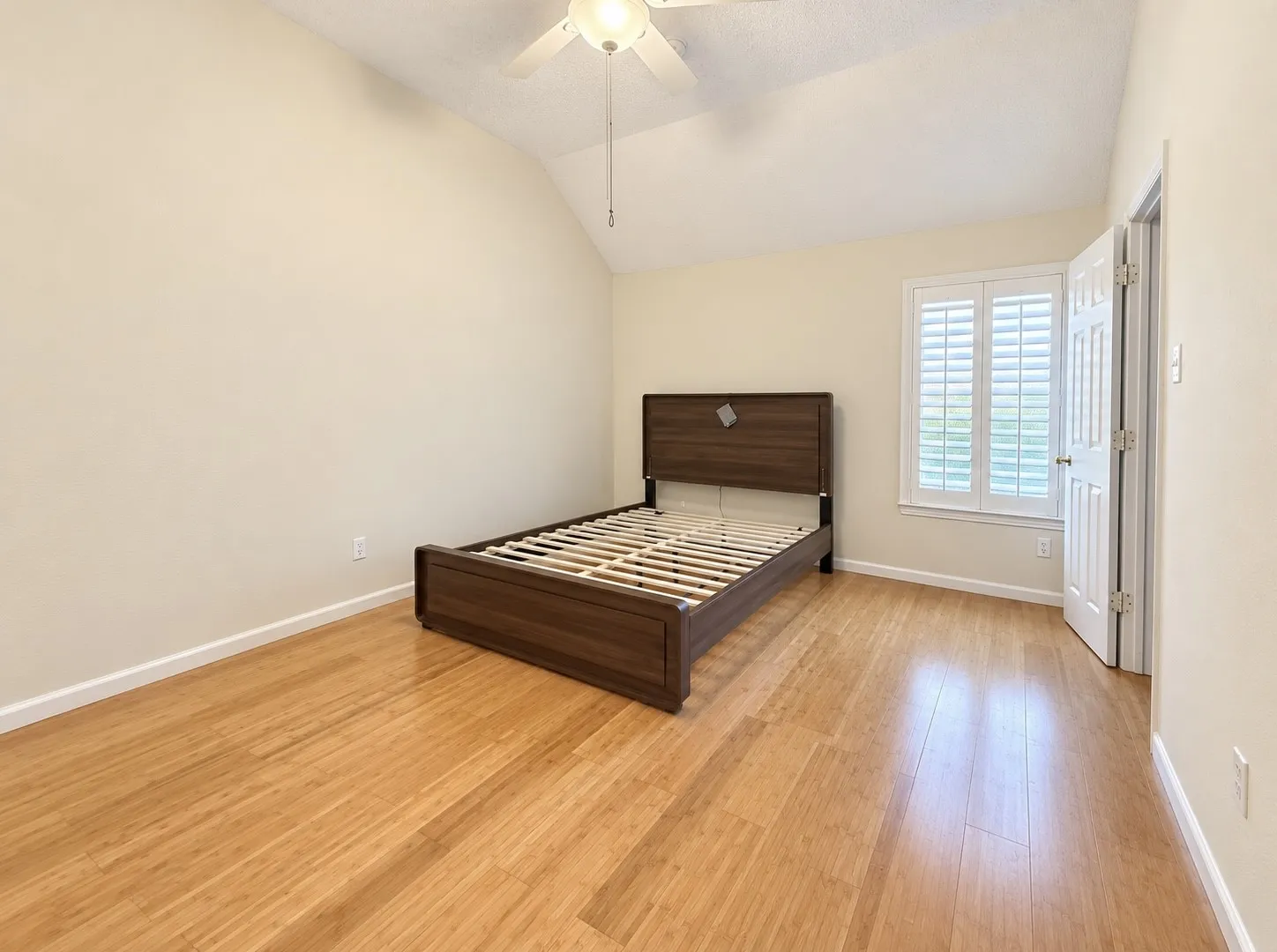 Light wood-finish flooring, vaulted ceiling, ceiling fan with integrated lighting, window with white plantation shutters, and a paneled interior door
