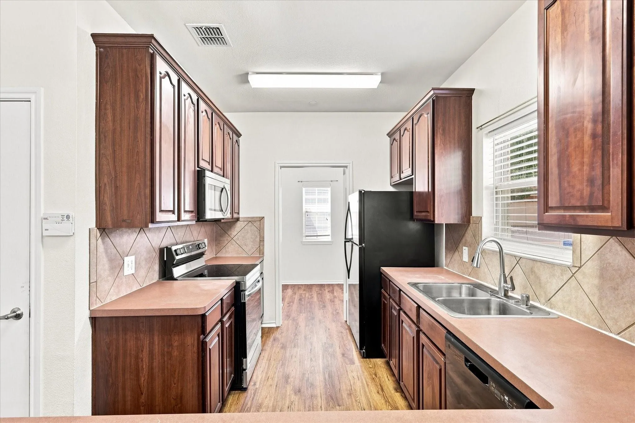 Galley kitchen featuring wood-finish cabinetry, tile backsplash, laminate countertops, and stainless steel sink