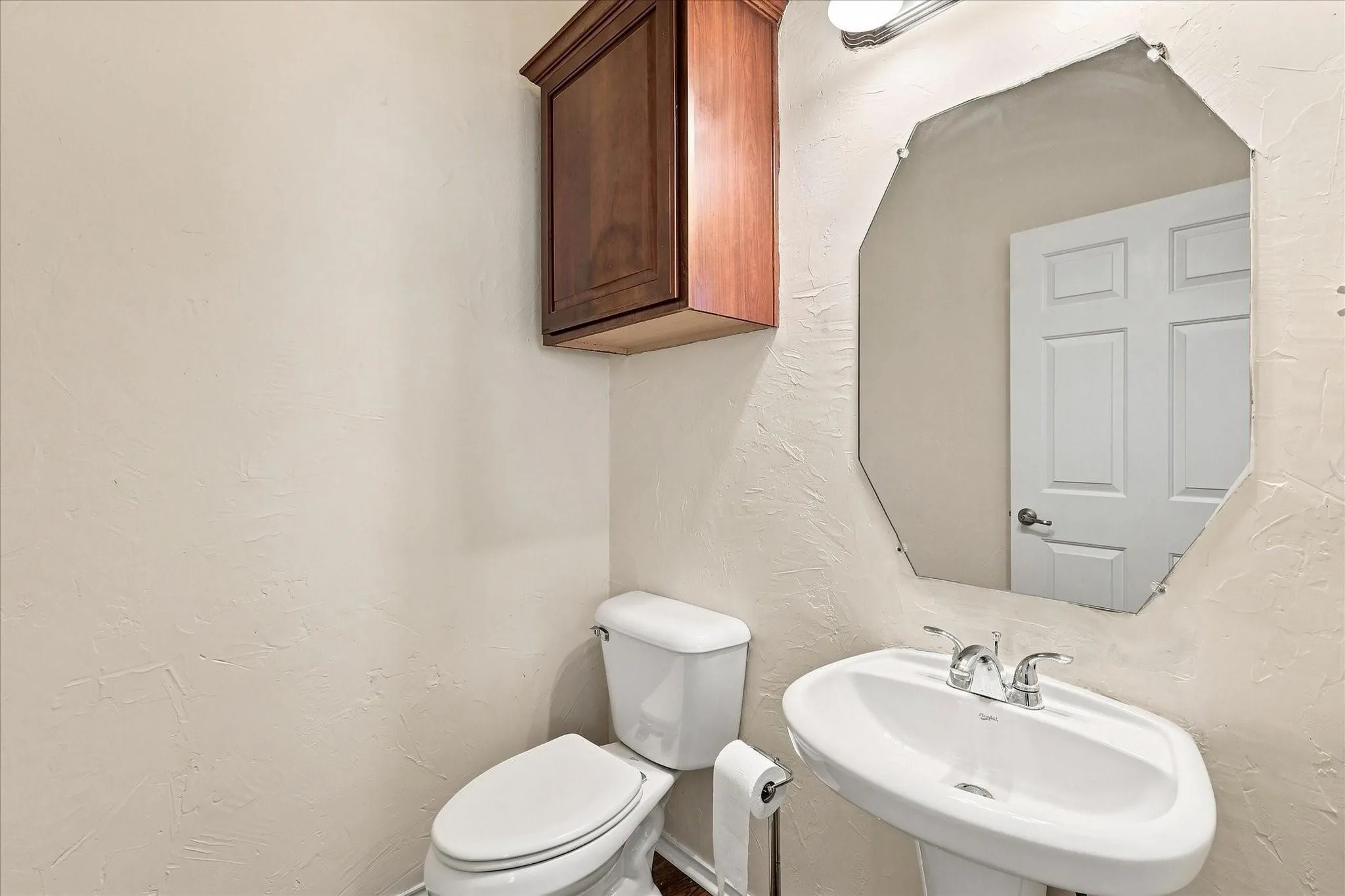 Pedestal sink with chrome fixtures, an octagonal wall-mounted mirror, and a wood-finish wall cabinet