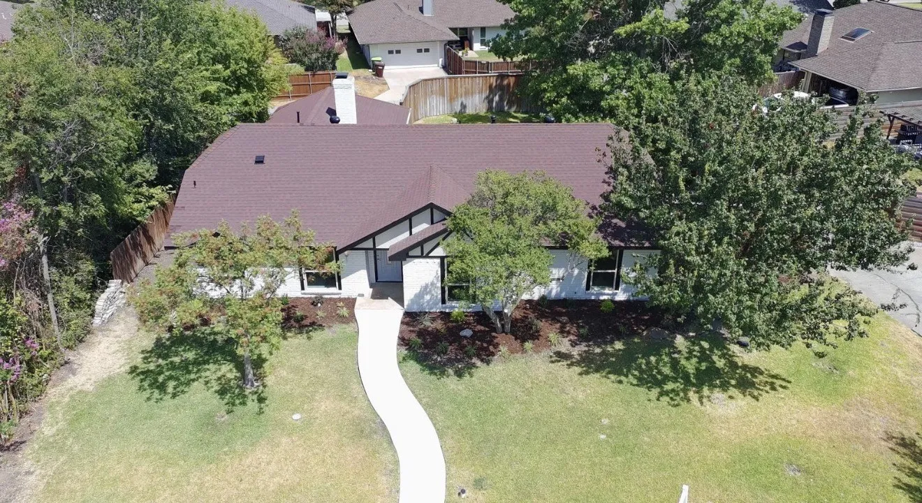 White exterior with dark trim accents, curved concrete walkway, mature trees, and a brown shingle roof