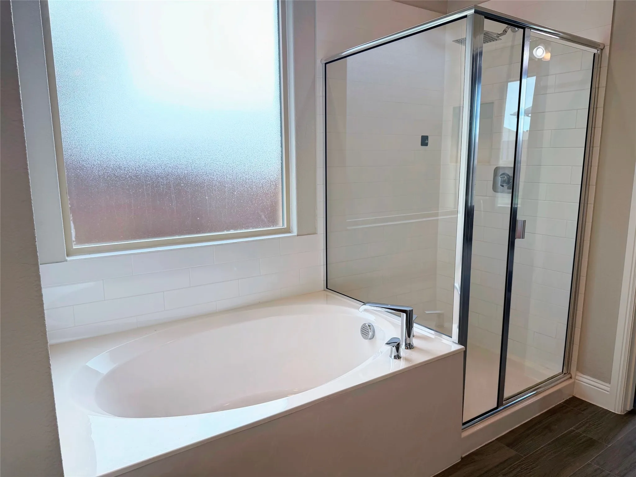 Built-in soaking tub with chrome fixtures, enclosed glass shower with subway tile surround, frosted window, and wood-finish flooring