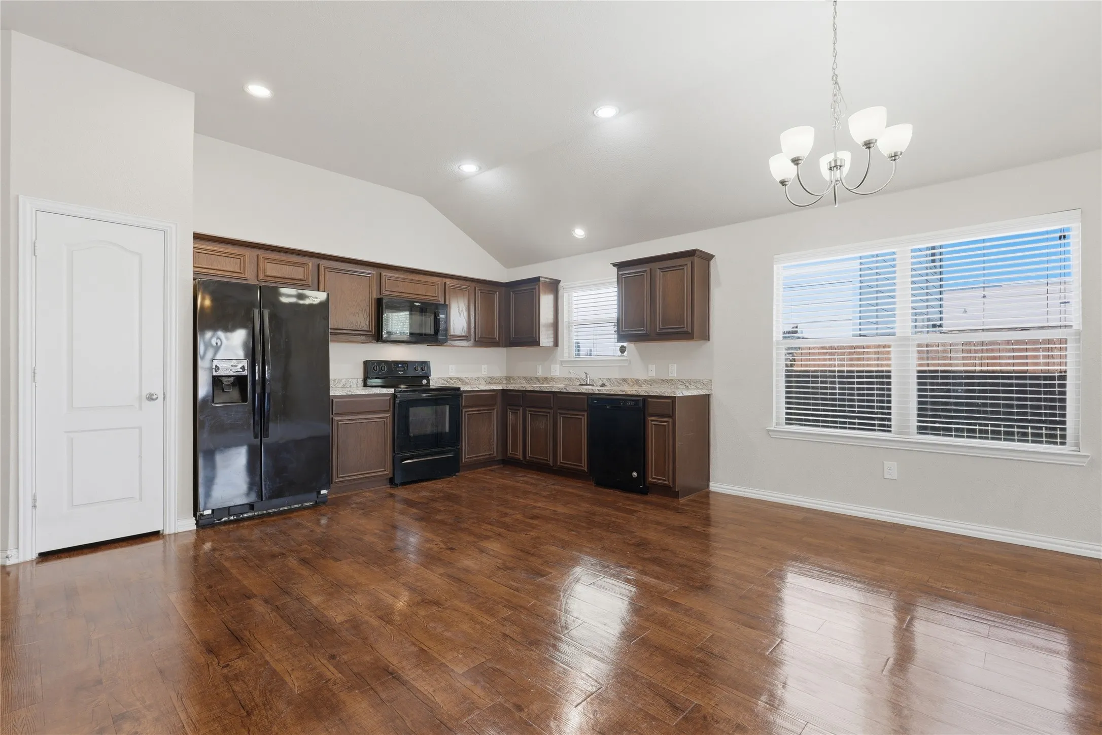 Open-concept kitchen and dining area featuring wood-finish flooring, vaulted ceilings, recessed lighting, brown cabinetry, and a five-light chandelier