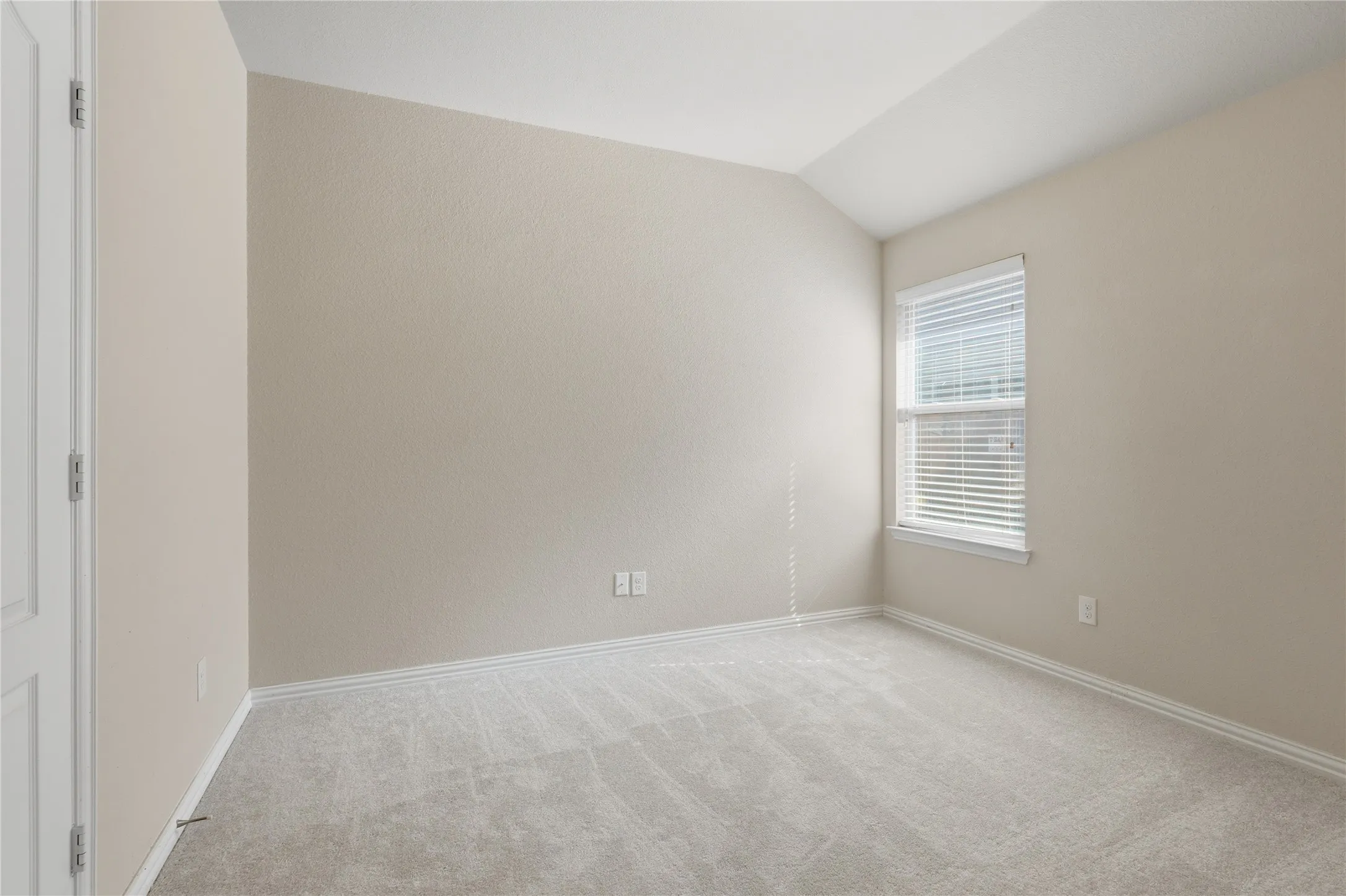 Vaulted ceiling, neutral wall tones, window with horizontal blinds, light-toned carpet, and a white interior door