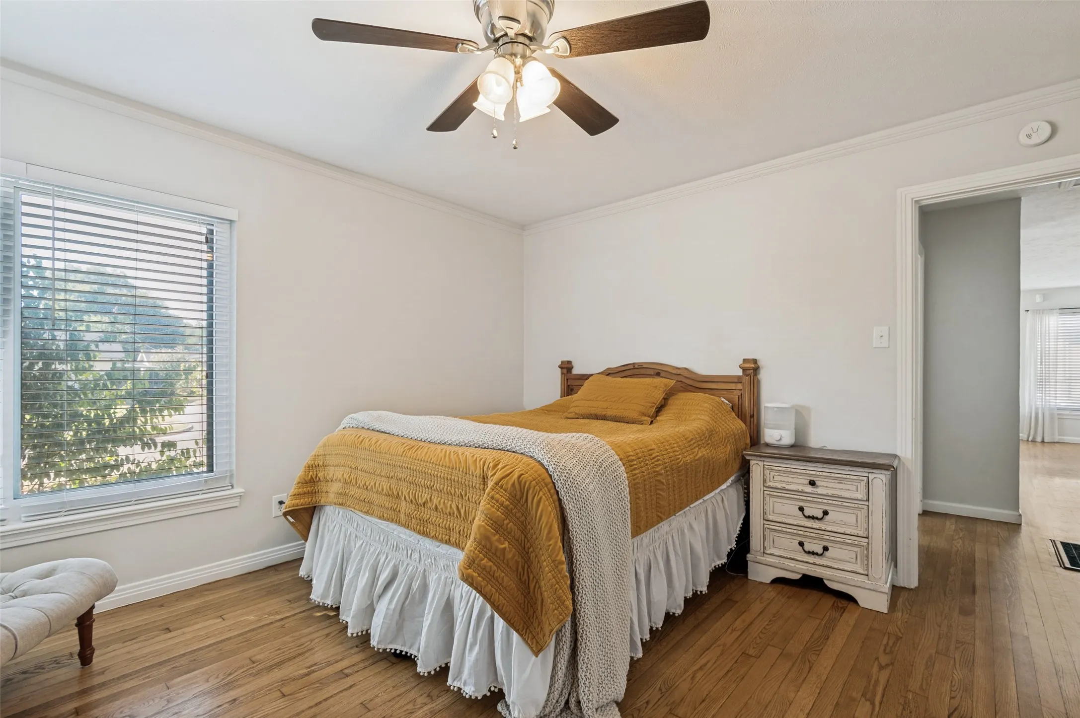 Hardwood flooring, ceiling fan with light fixture, white interior walls, crown molding, and window with blinds