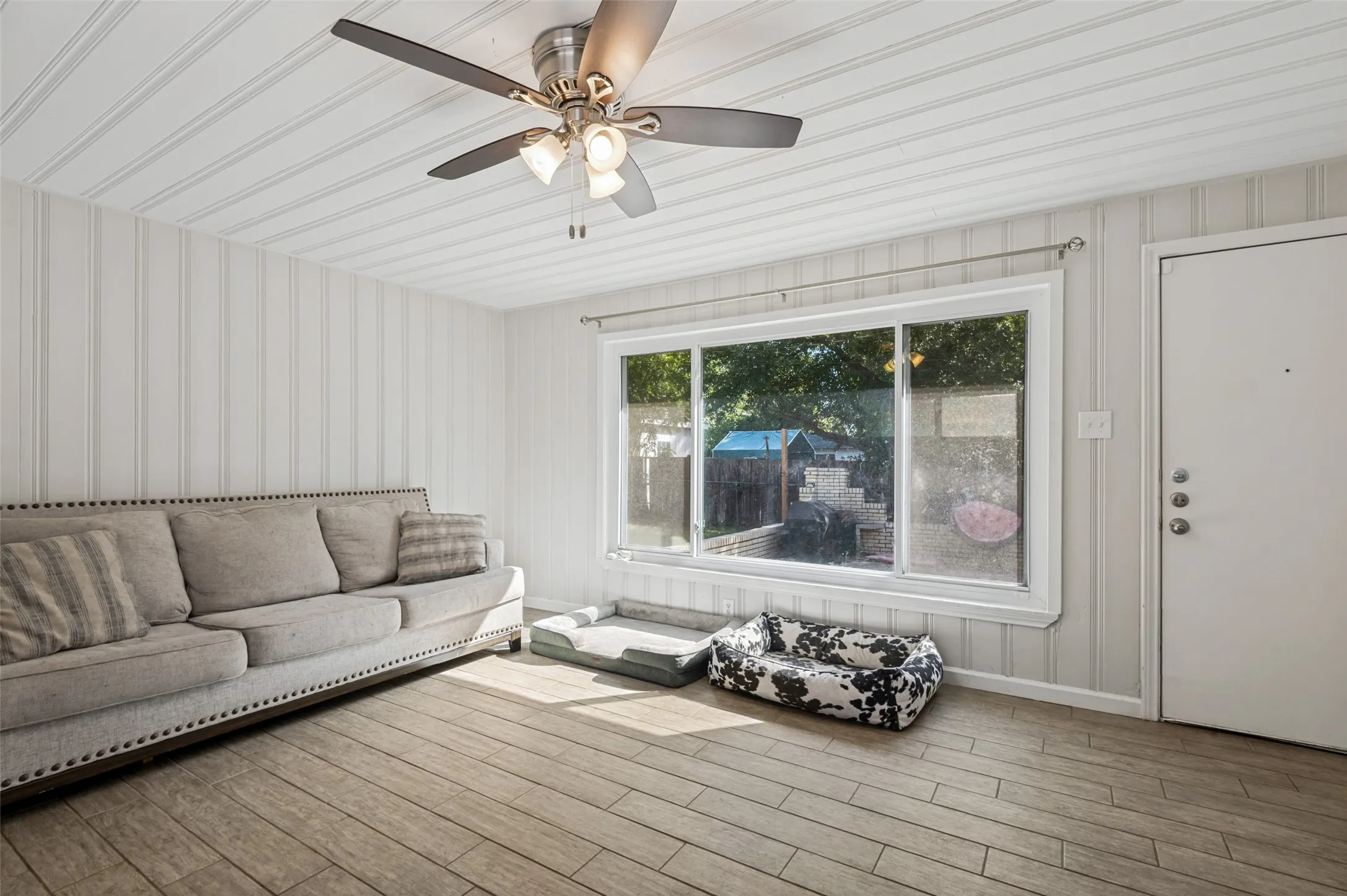 Wood-finish flooring, ceiling fan with integrated lighting, paneled walls, and a large window