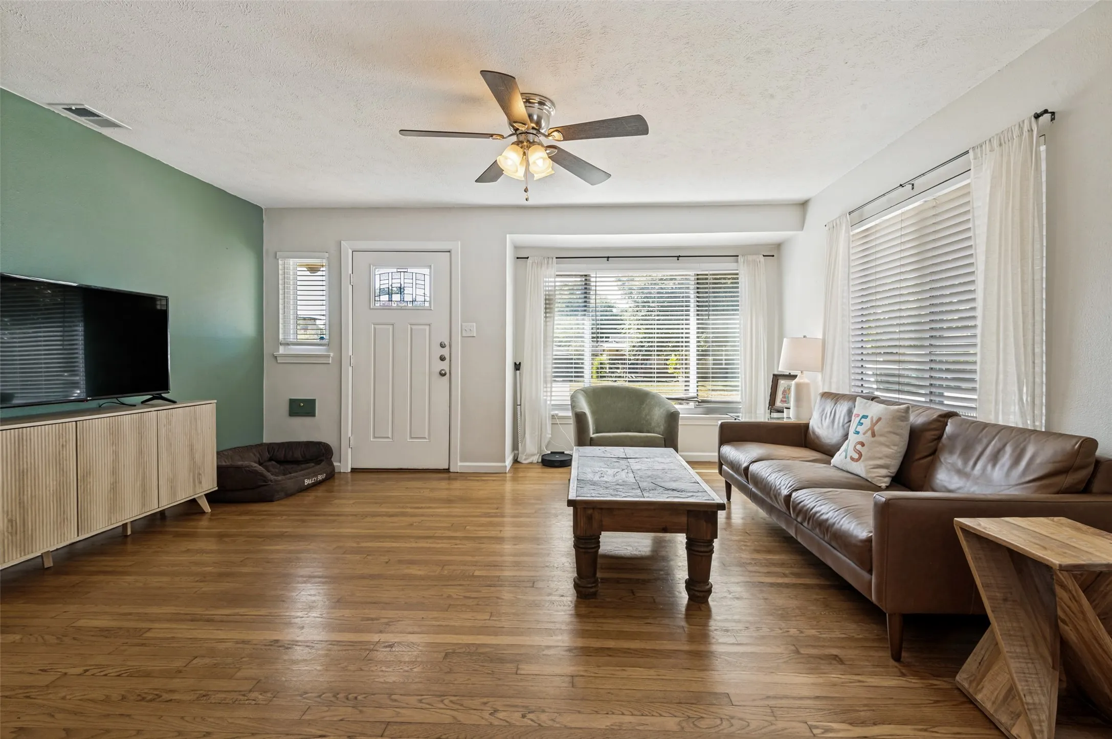 Hardwood flooring, painted accent wall, ceiling fan, front entry door with transom window, and large bay window