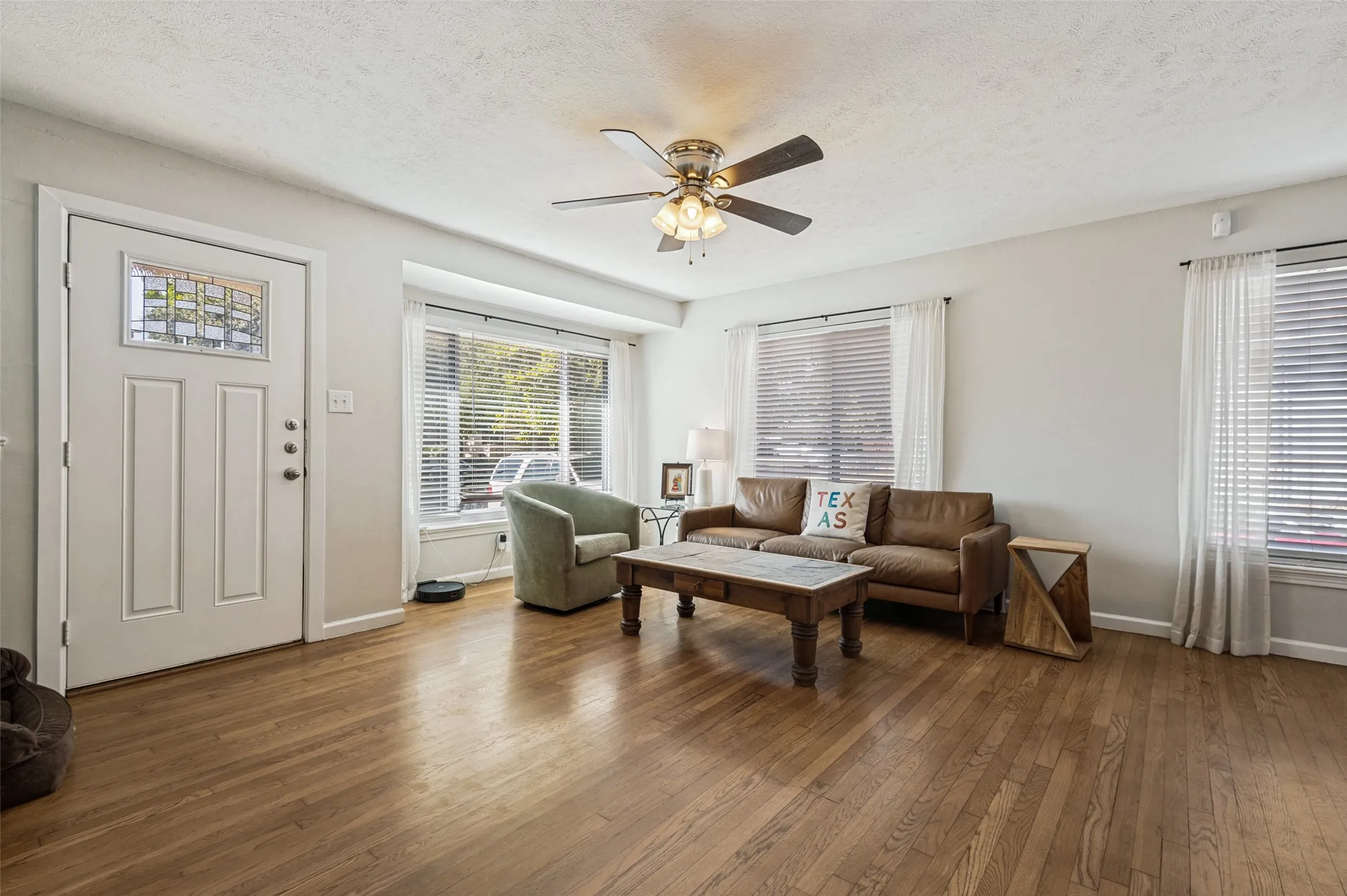 Wood-finish flooring throughout, multiple windows with blinds, ceiling fan, and a paneled entry door with decorative glass