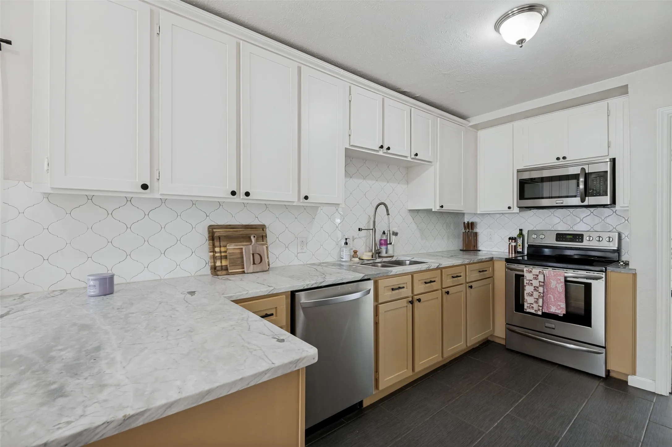White shaker-style upper cabinetry with black hardware, light wood-finish lower cabinetry, light-toned countertops, stainless steel appliances, and dark rectangular floor tiling