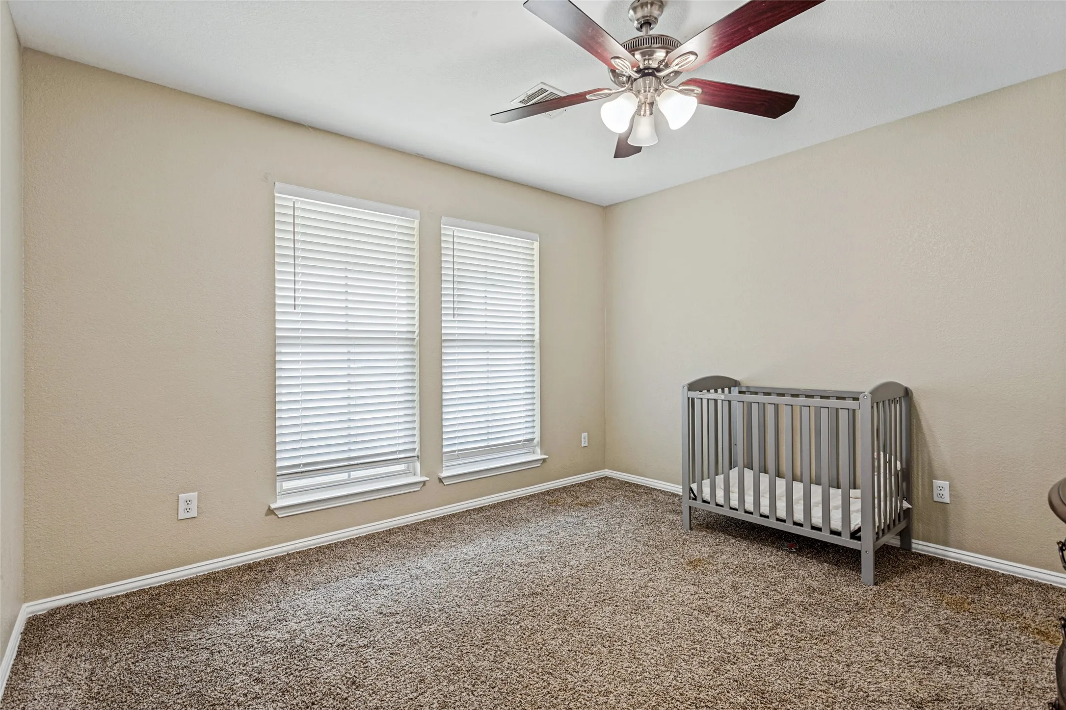 Neutral wall paint, plush carpet flooring, two windows with blinds, and a ceiling fan with integrated lighting