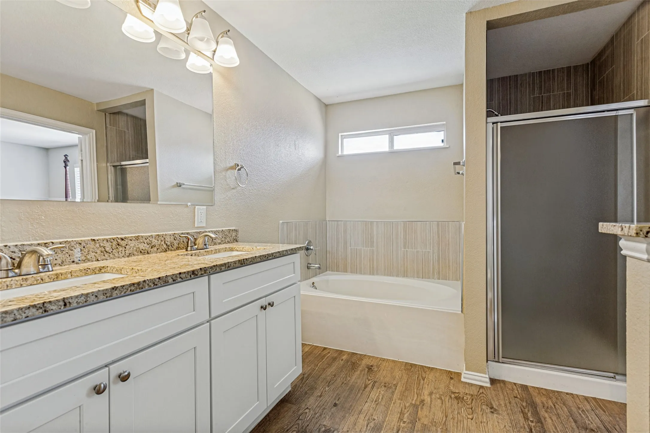 Dual vanity with granite-style countertop, wood-finish flooring, built-in bathtub, and a separate shower with frosted glass enclosure