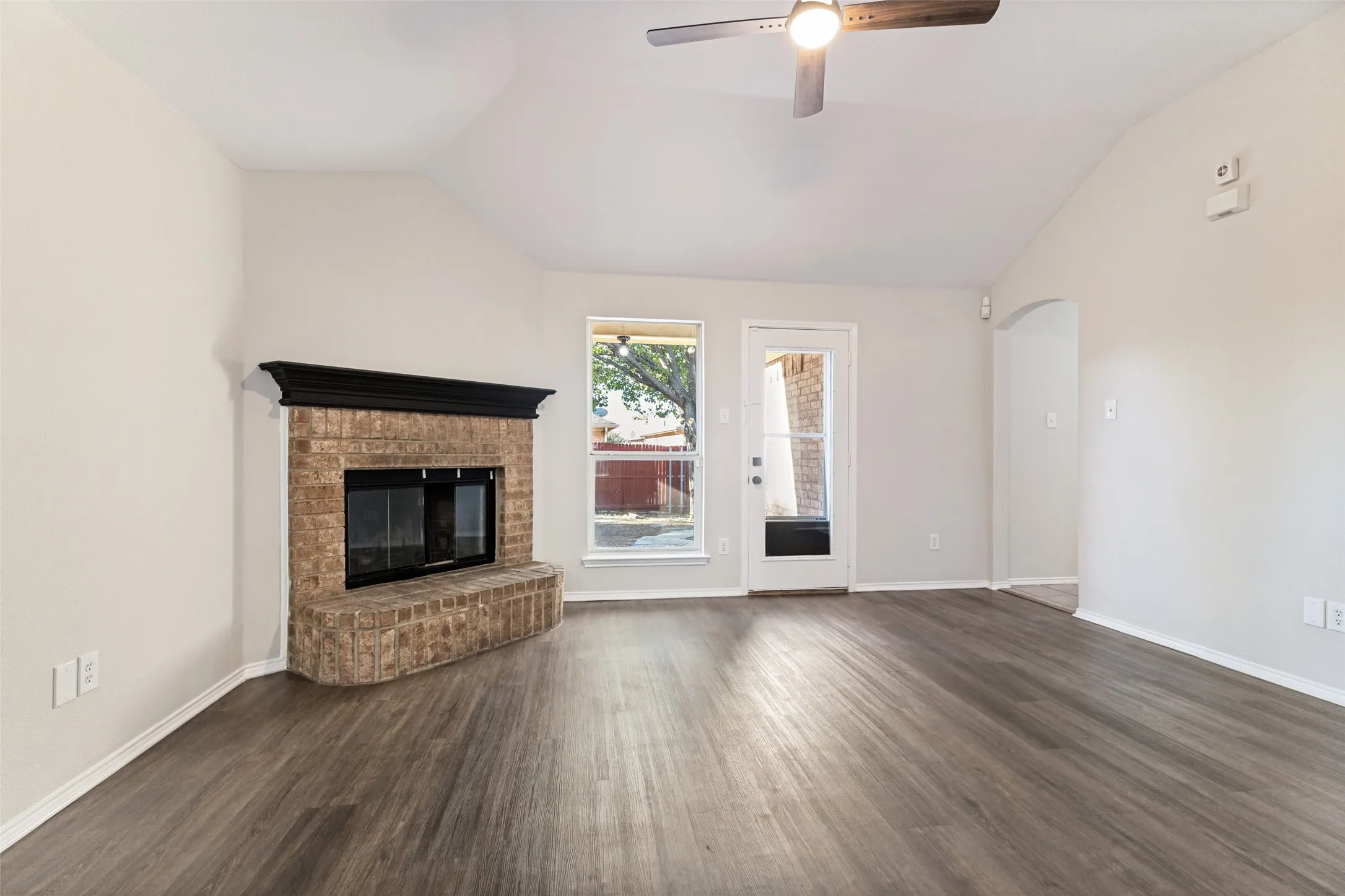 Vaulted ceiling with a ceiling fan, brick fireplace with a dark wood-finish mantel, wood-finish flooring, and a glass-paneled door