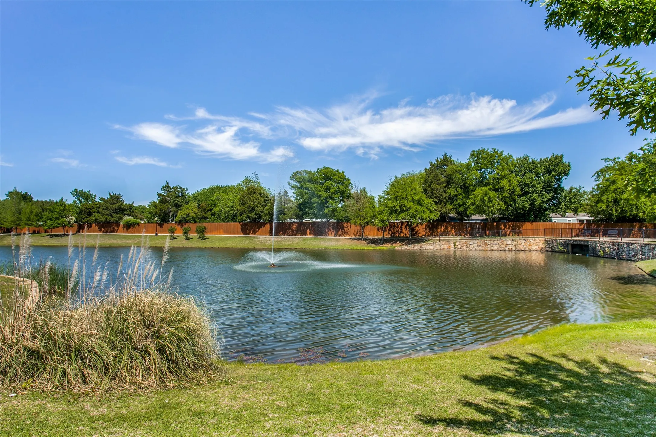 Pond with a central water feature, surrounded by a manicured lawn, established landscaping, and a perimeter fence