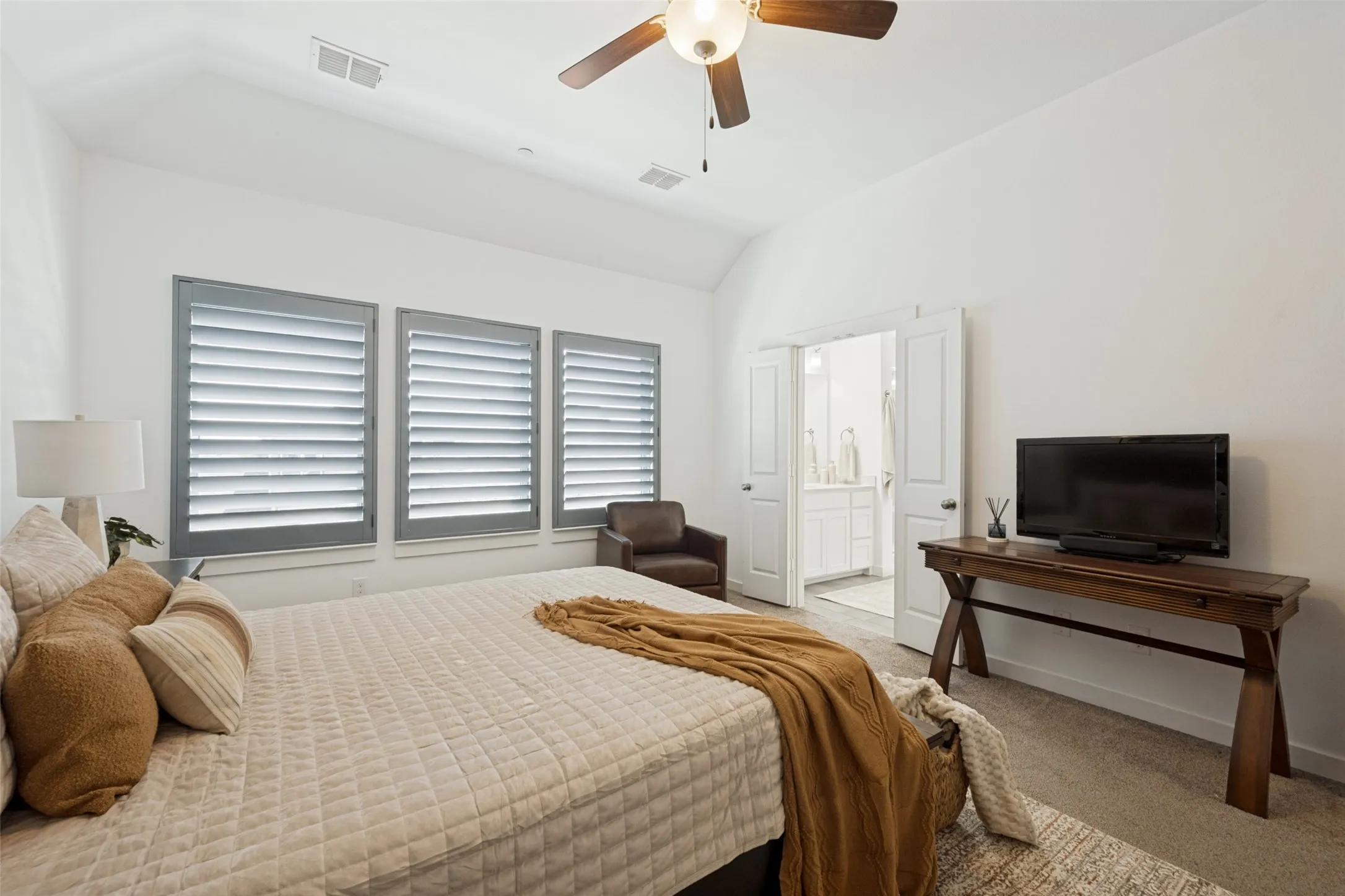 Vaulted ceiling with an attached ceiling fan, three windows with louvered shutters, and double doors opening to an adjacent bathroom featuring a dual vanity