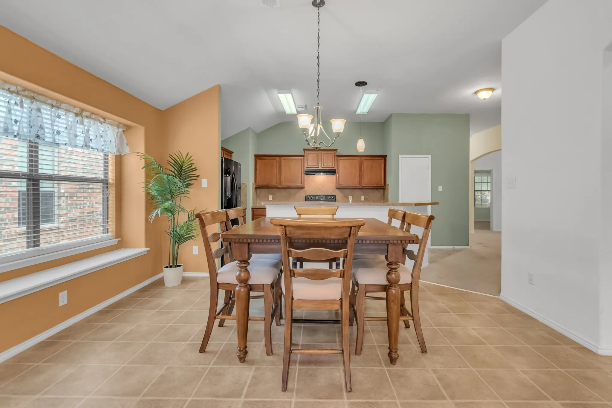 Open dining and kitchen area featuring warm wood-finish cabinetry, neutral tile flooring, large windows with natural light, pendant lighting, and ample counter space for meal preparation and entertaining