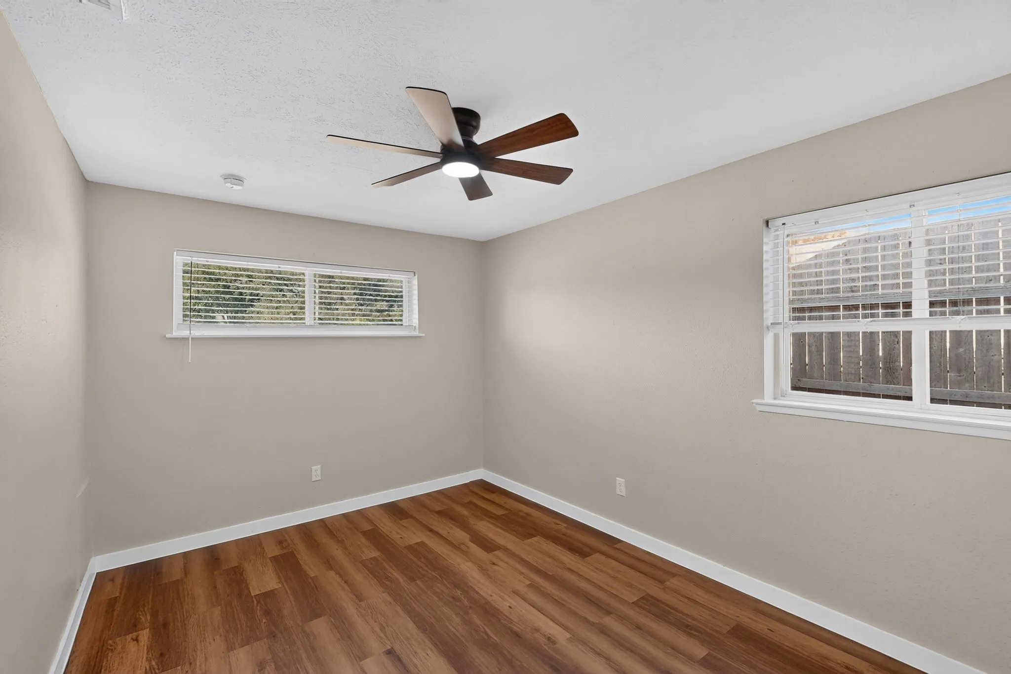 Wood-finish flooring, neutral wall tones, two windows with blinds, and a ceiling fan with integrated lighting