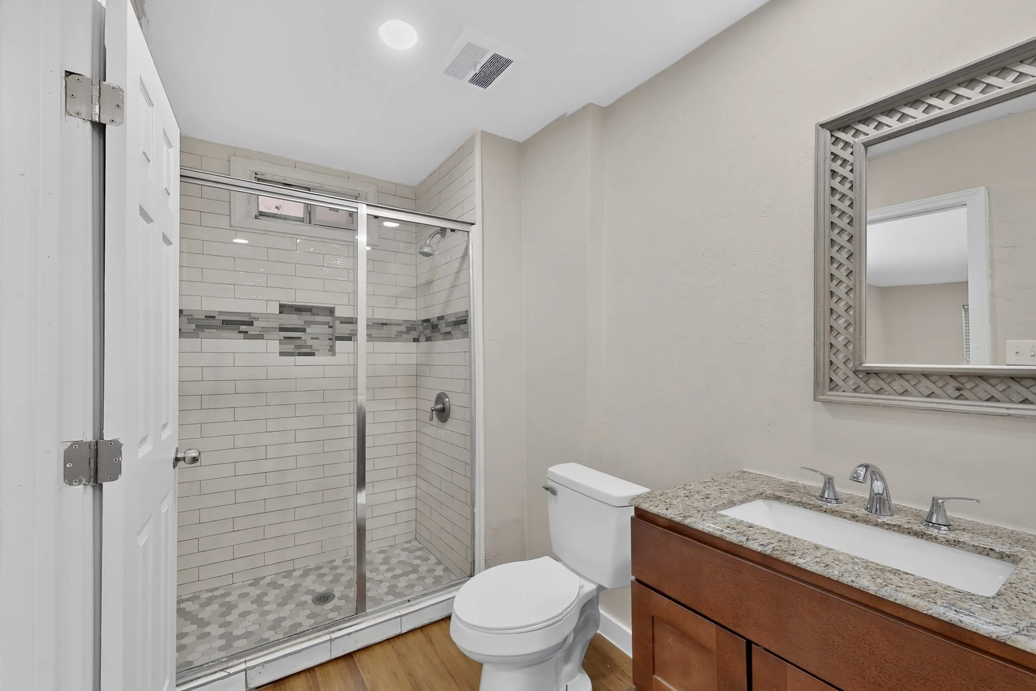 Walk-in shower with subway tile and glass enclosure, wood-finish flooring, vanity with granite countertop, and framed mirror
