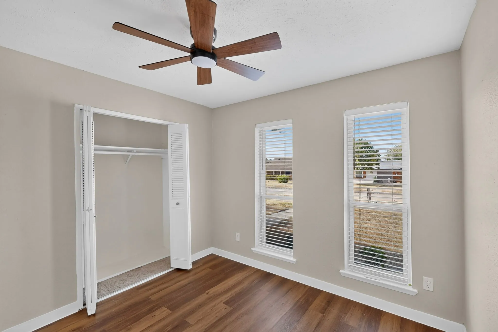 Wood-finish flooring, neutral wall paint, two windows with blinds, bi-fold closet doors, and a ceiling fan with integrated lighting