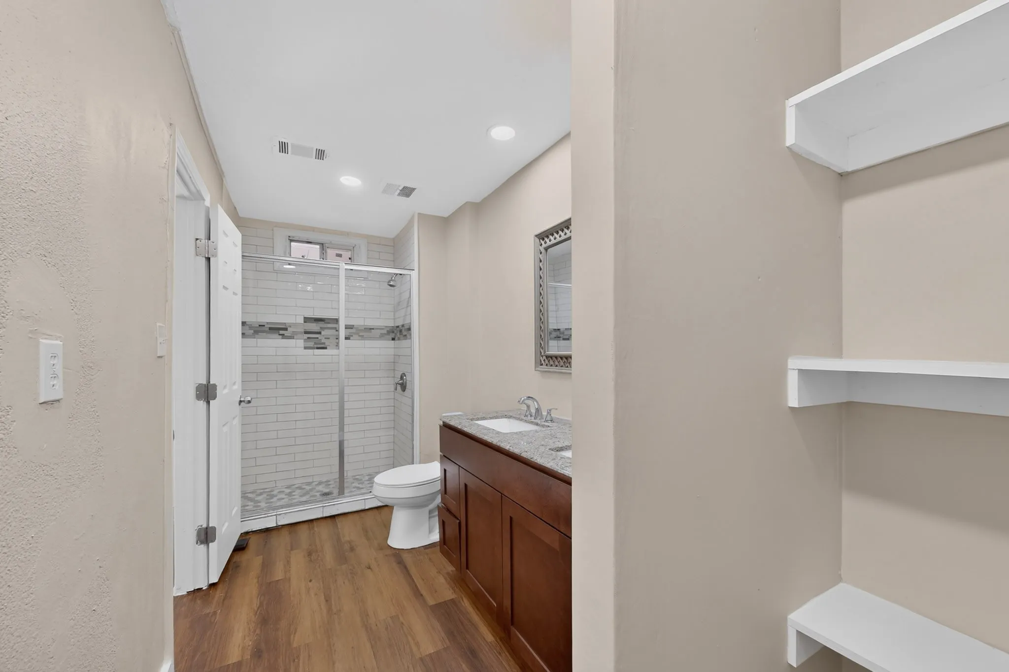 Glass-enclosed shower with subway tile surround, wood-finish flooring, dual-sink vanity with light-toned countertop, and built-in shelving