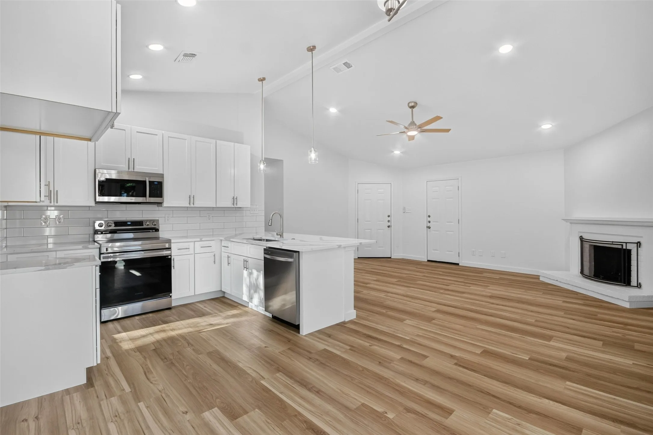 Open-concept living area featuring wood-finish flooring, a contemporary kitchen with stainless steel appliances, white cabinetry, and an island with pendant lighting, and a corner fireplace