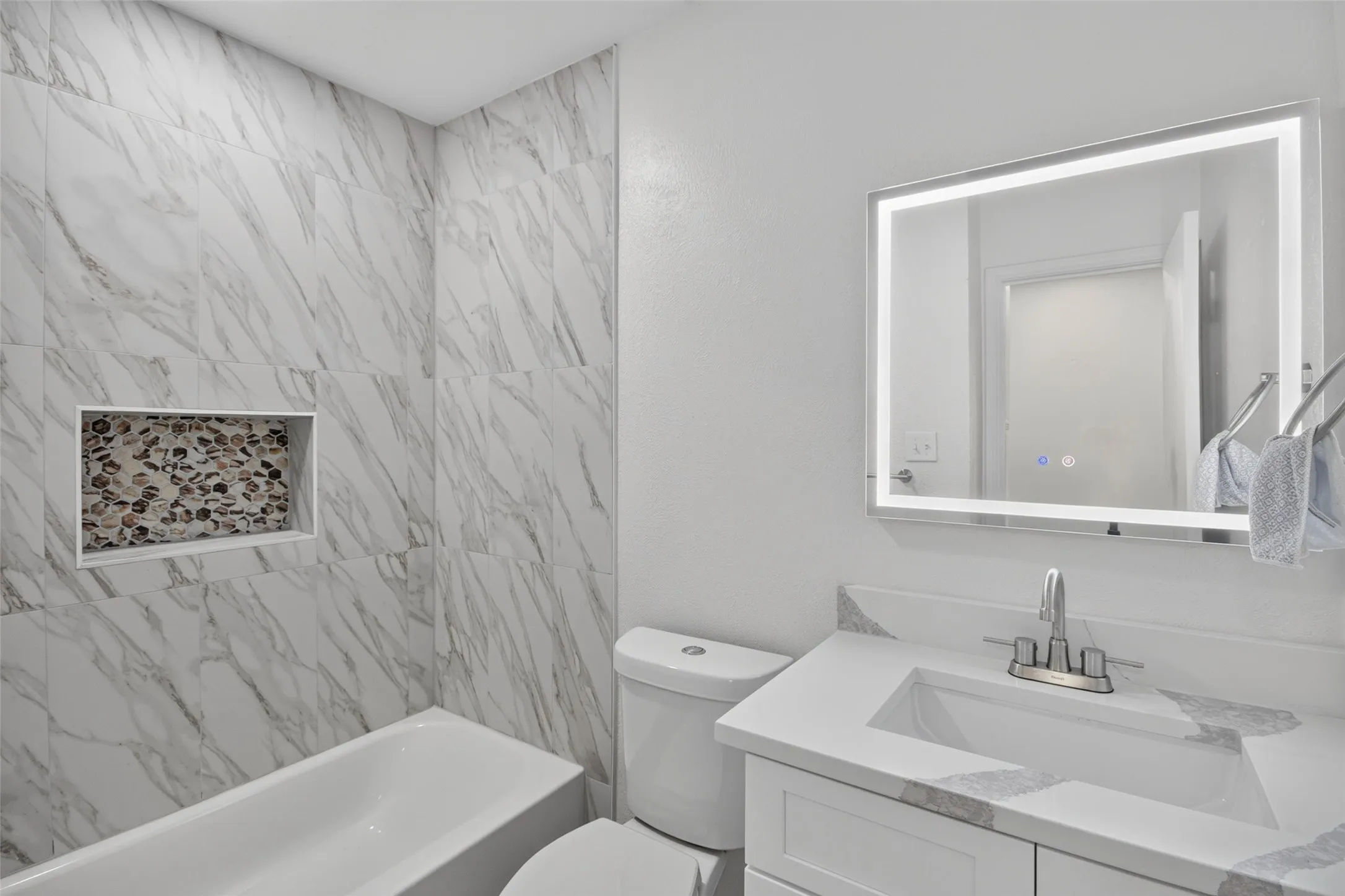 Bathroom featuring a built-in tub, marble-look wall tile, a stone-tiled shower niche, a vanity with an integrated sink, and an illuminated mirror