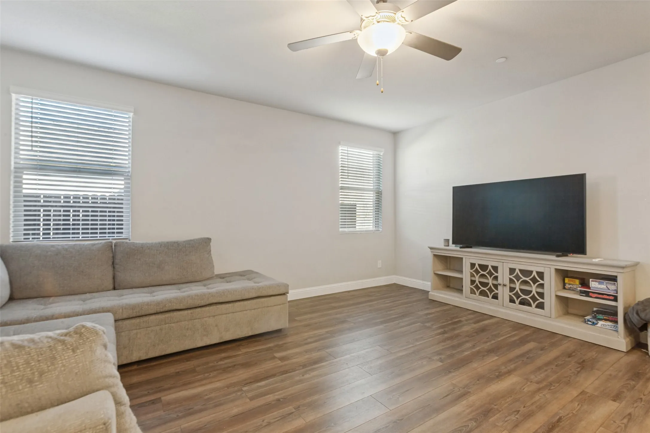 Spacious living room showcasing wood-finish flooring, abundant natural light from dual windows with plantation shutters, ceiling fan with integrated lighting, and a built-in media console with open shelving and decorative lattice-front cabinetry