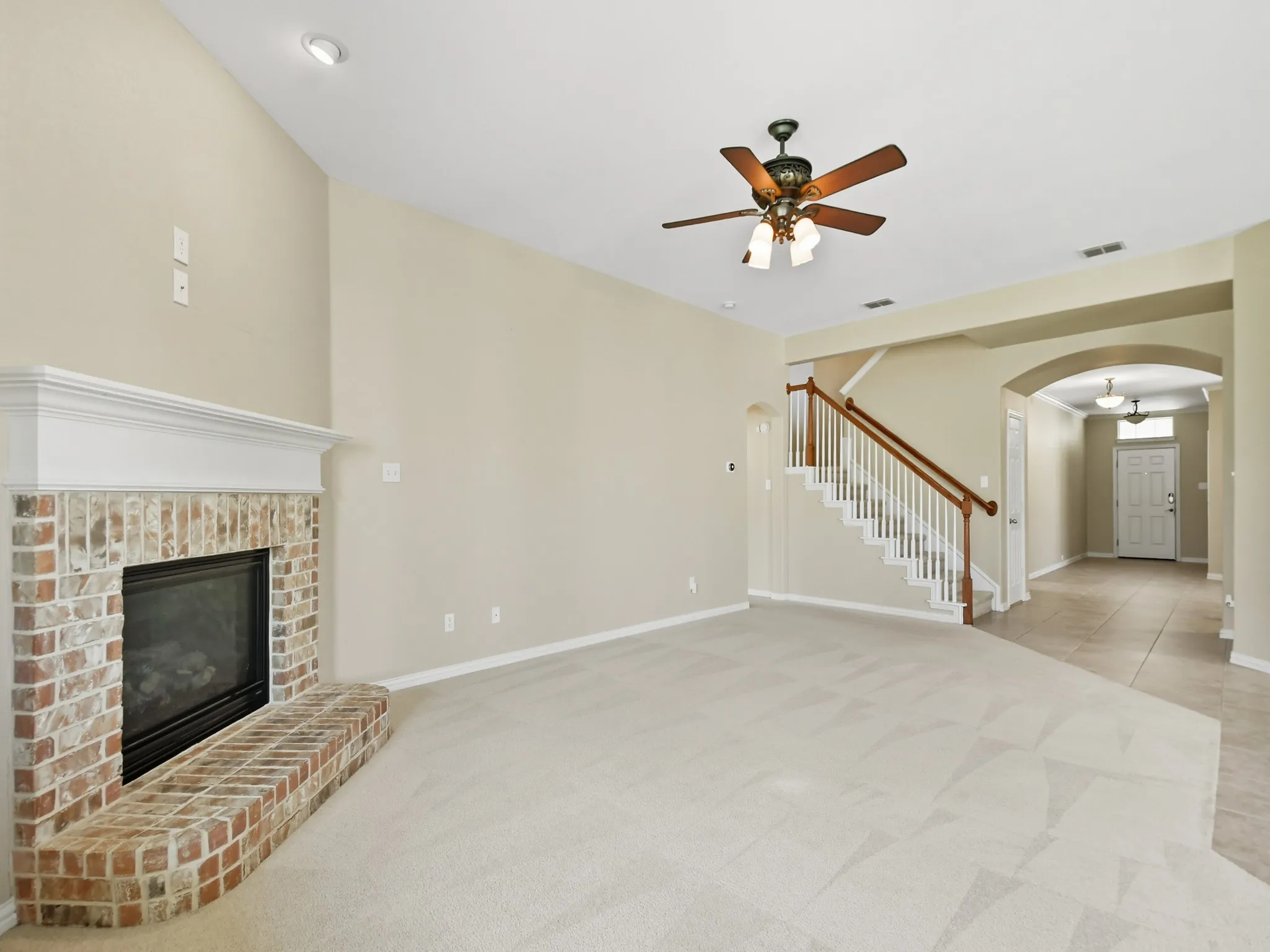Brick fireplace with white mantel, vaulted ceiling with recessed lighting, ceiling fan with light fixture, wood and white spindle staircase, and tiled entryway with arched doorway