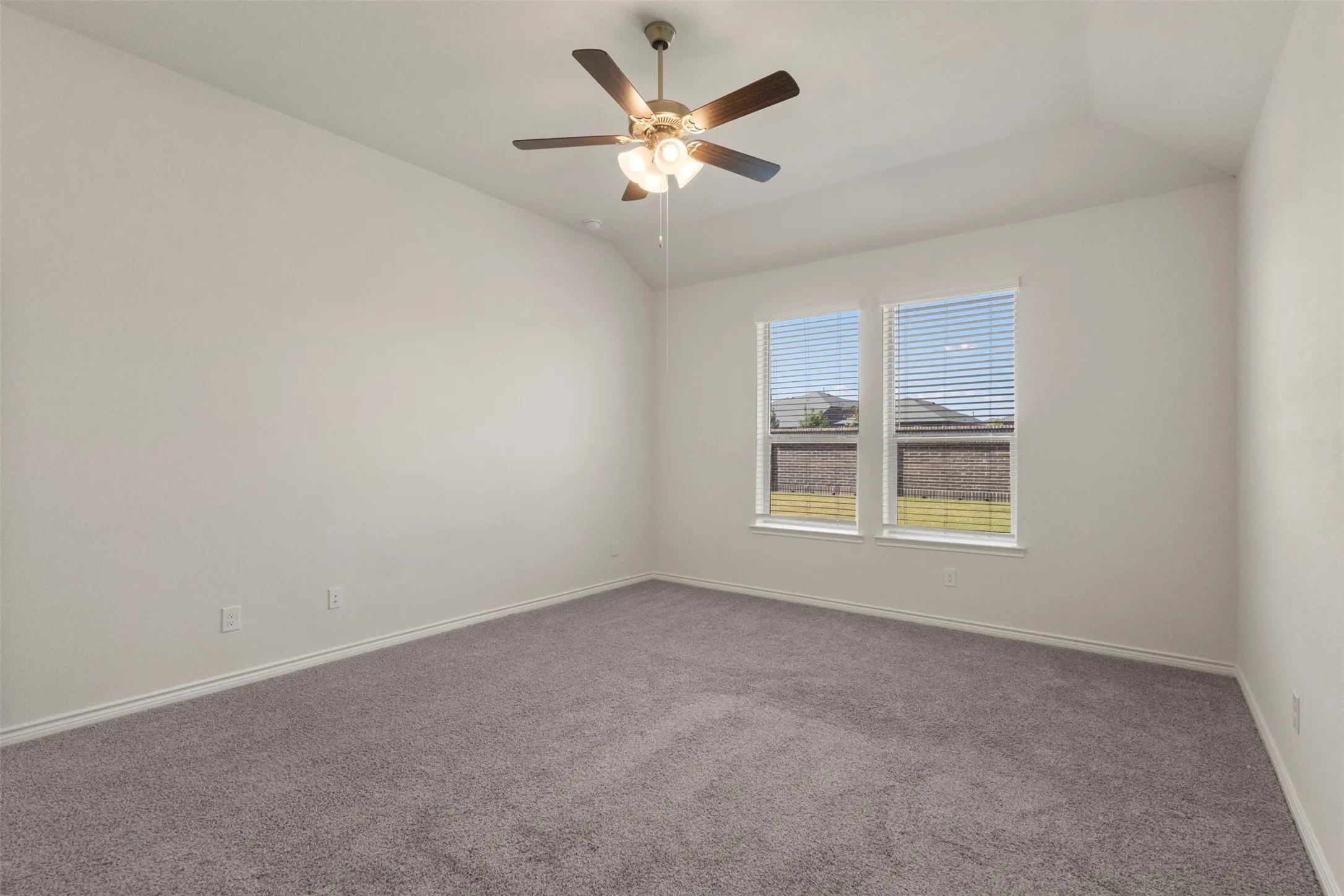 Vaulted ceiling, ceiling fan with light fixture, two windows with blinds, neutral carpet, and white wall paint