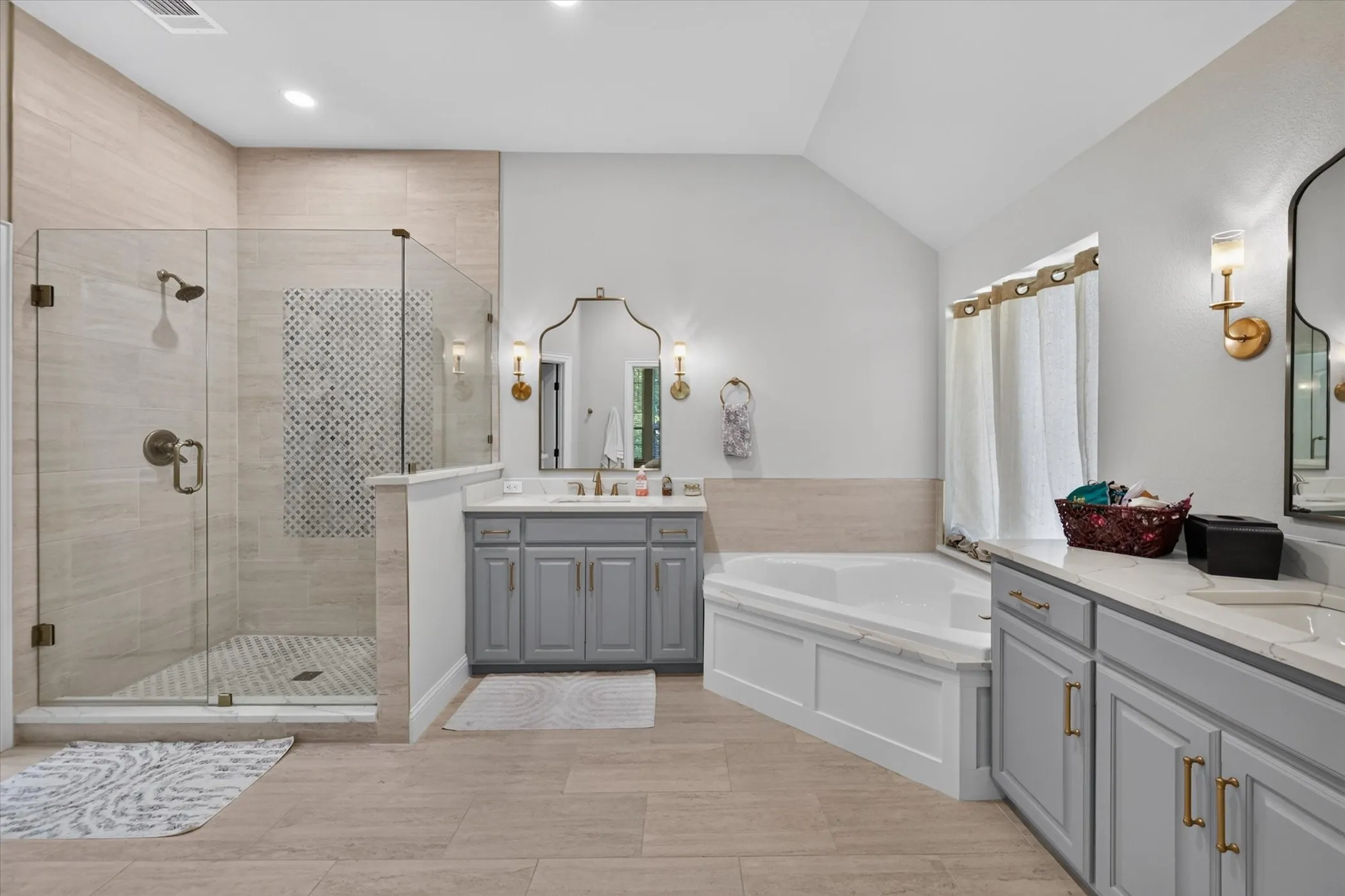 Spacious bathroom featuring a glass-enclosed shower with tile surround, a built-in corner bathtub, dual vanities with light gray cabinetry, and sconce lighting