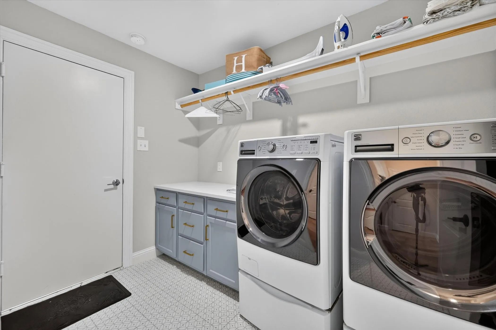 Laundry area with white patterned tile flooring, built-in lower cabinetry with gold-tone hardware, a white countertop, and an upper shelving system