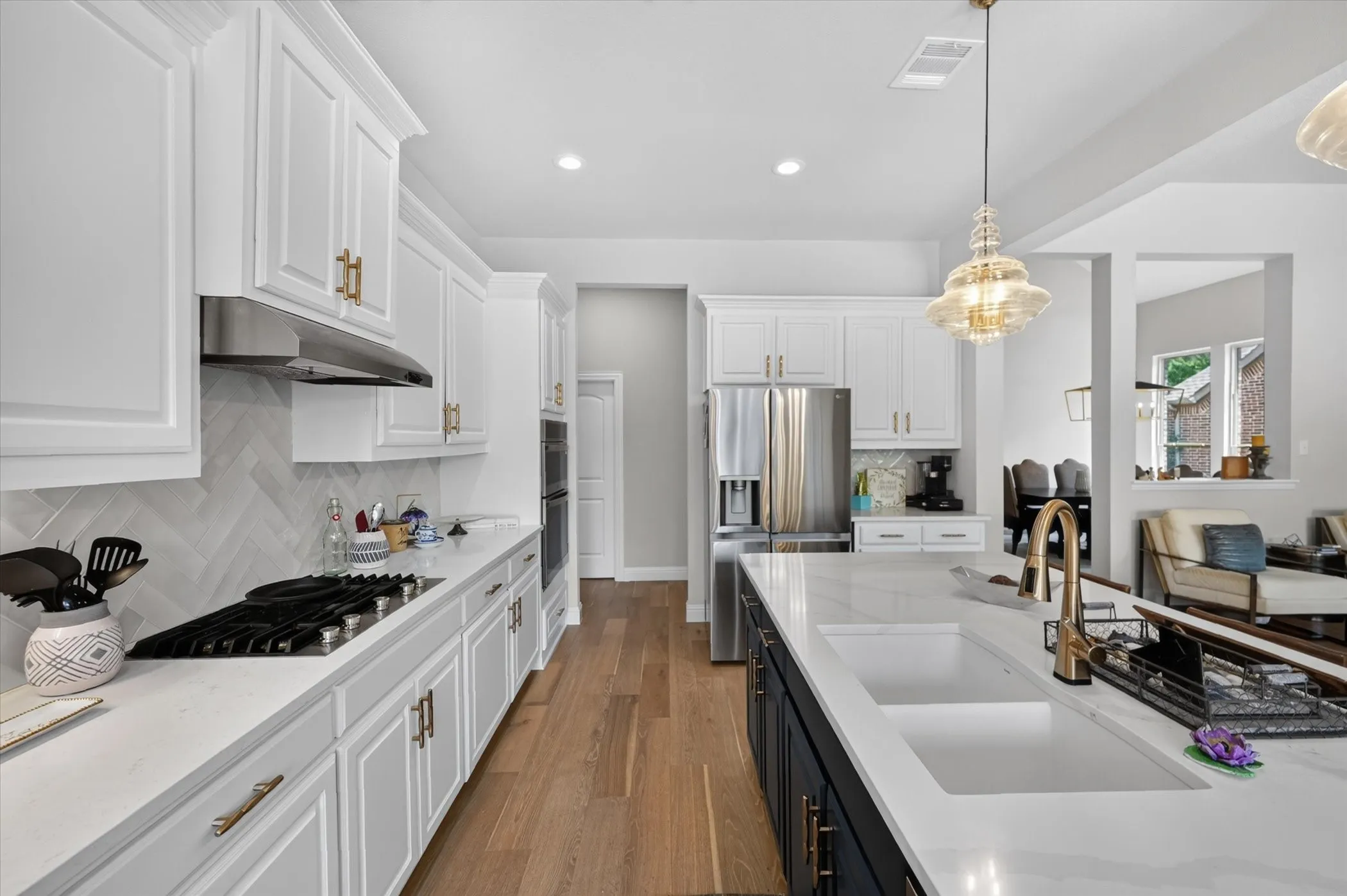 White cabinetry with gold-finish hardware, herringbone tile backsplash, stainless steel appliances, central island with double sink, and wood-finish flooring