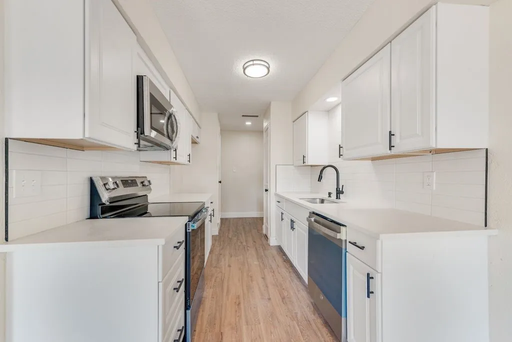 Galley kitchen featuring white cabinetry, light countertops, wood-finish flooring, stainless steel appliances, and a tile backsplash