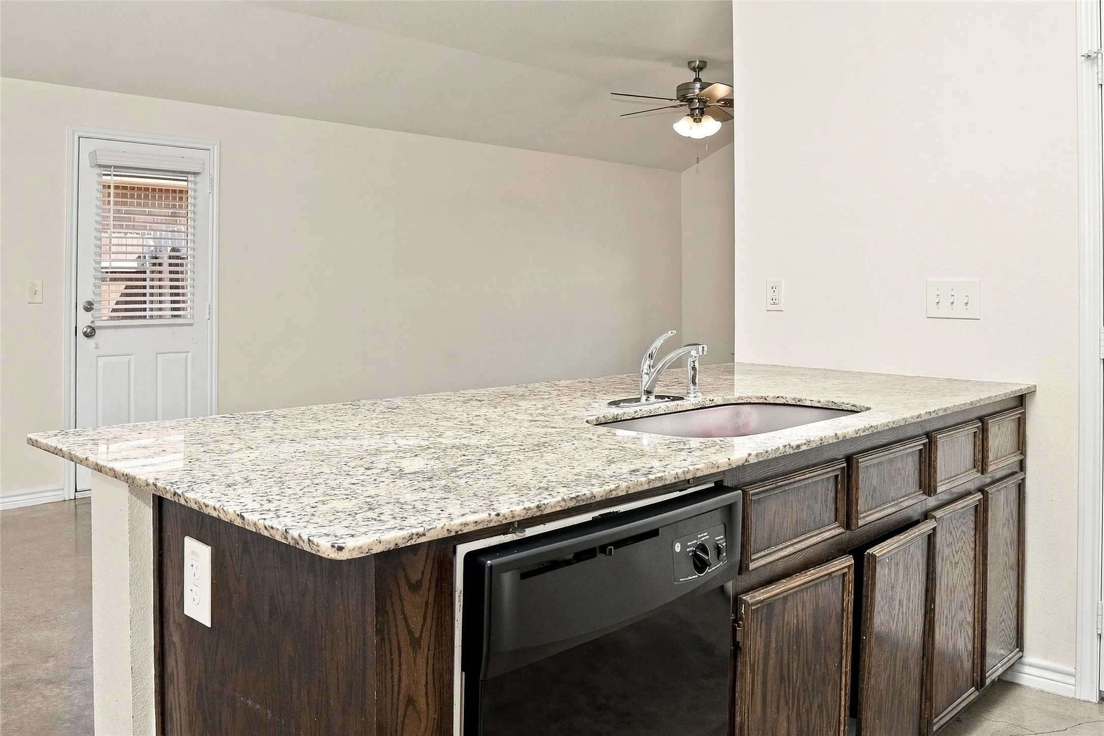 Kitchen island with granite countertop, stainless steel sink, dark wood-finish cabinetry, and built-in dishwasher