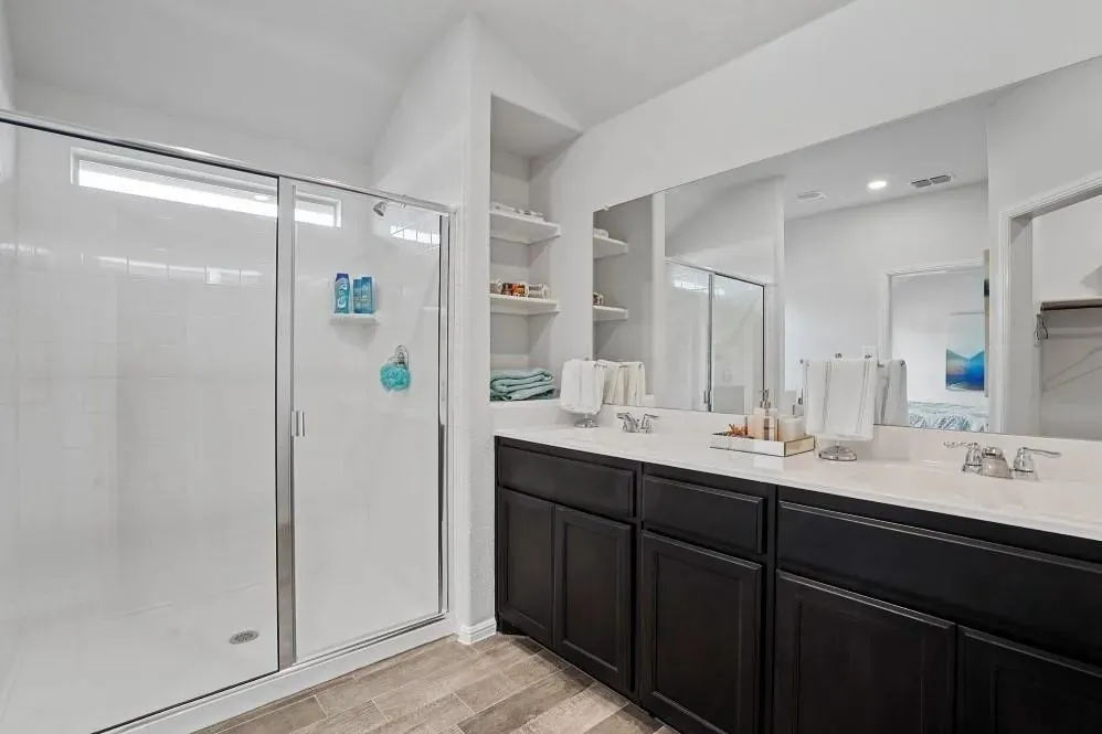 Dual vanity with white countertops and dark cabinetry, glass-enclosed shower with white subway tile, wood-finish flooring, and recessed shelving