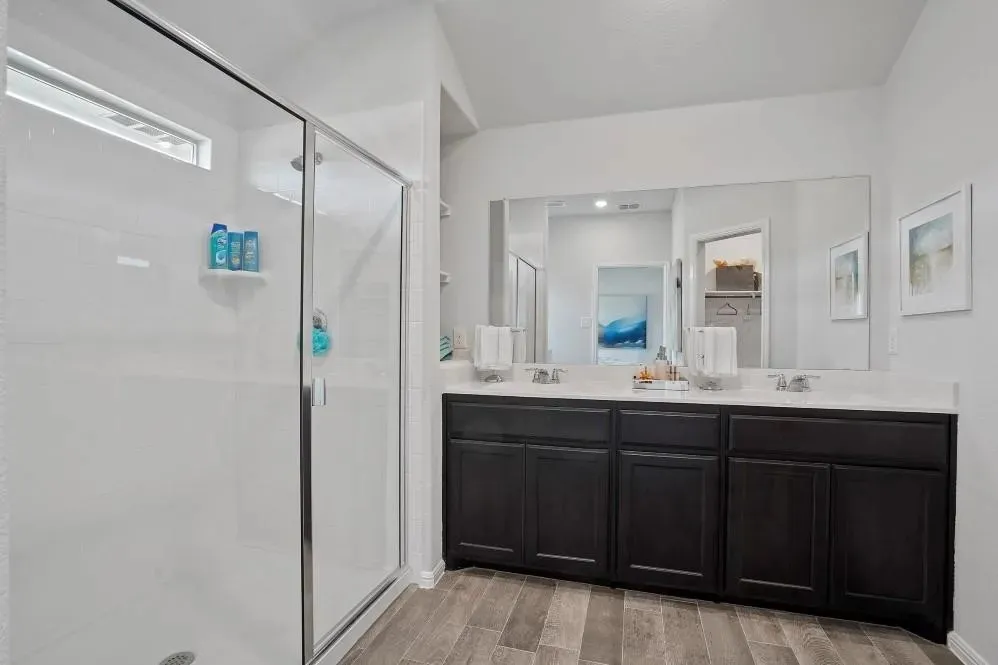 Walk-in shower with clear glass enclosure, dual vanity with dark wood-finish cabinetry, white countertop, and wood-finish flooring