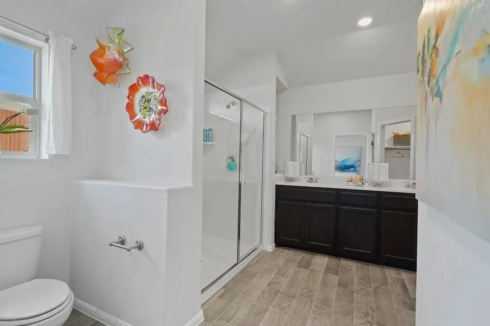 Dual sink vanity with dark wood-finish cabinetry, a glass-enclosed shower, wood-finish flooring, and a toilet