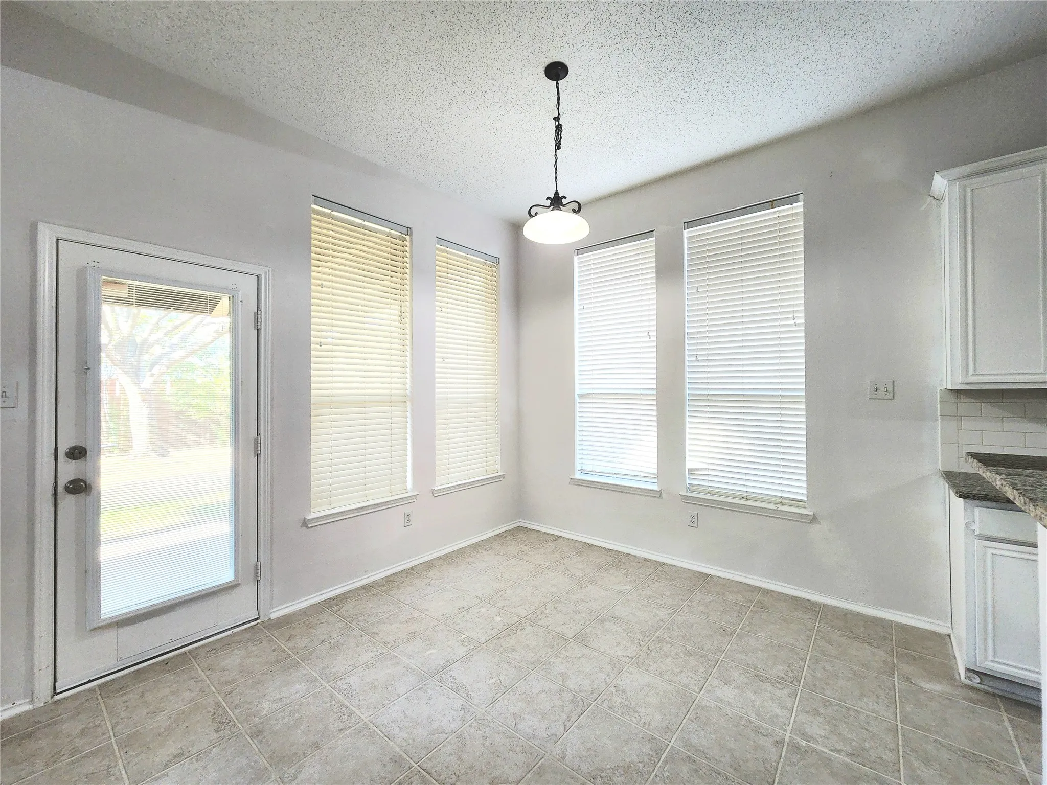Tile flooring, multiple windows with blinds, white cabinetry, light-colored backsplash, and a pendant light fixture