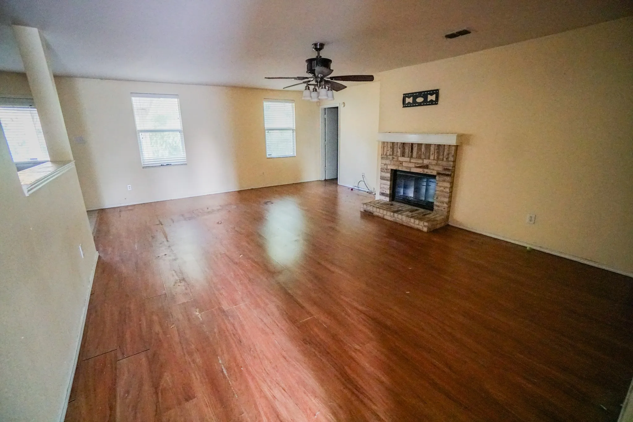 Spacious interior featuring wood-finish flooring, a brick-surround fireplace with a white mantel, multiple windows, and a ceiling fan with integrated lighting