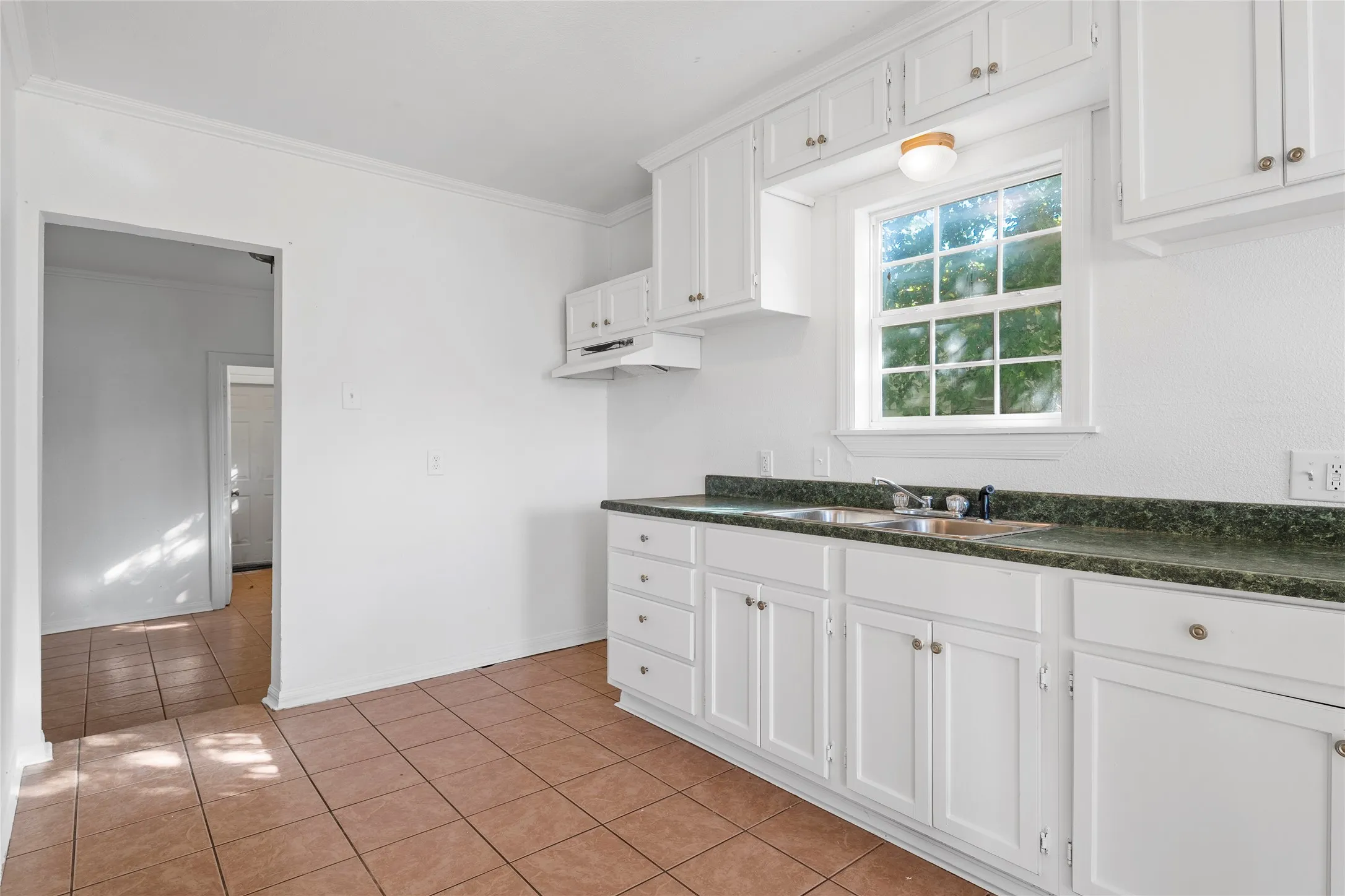 Kitchen with dark countertops, white cabinetry, ornamental molding, light tile patterned floors, and under cabinet range hood