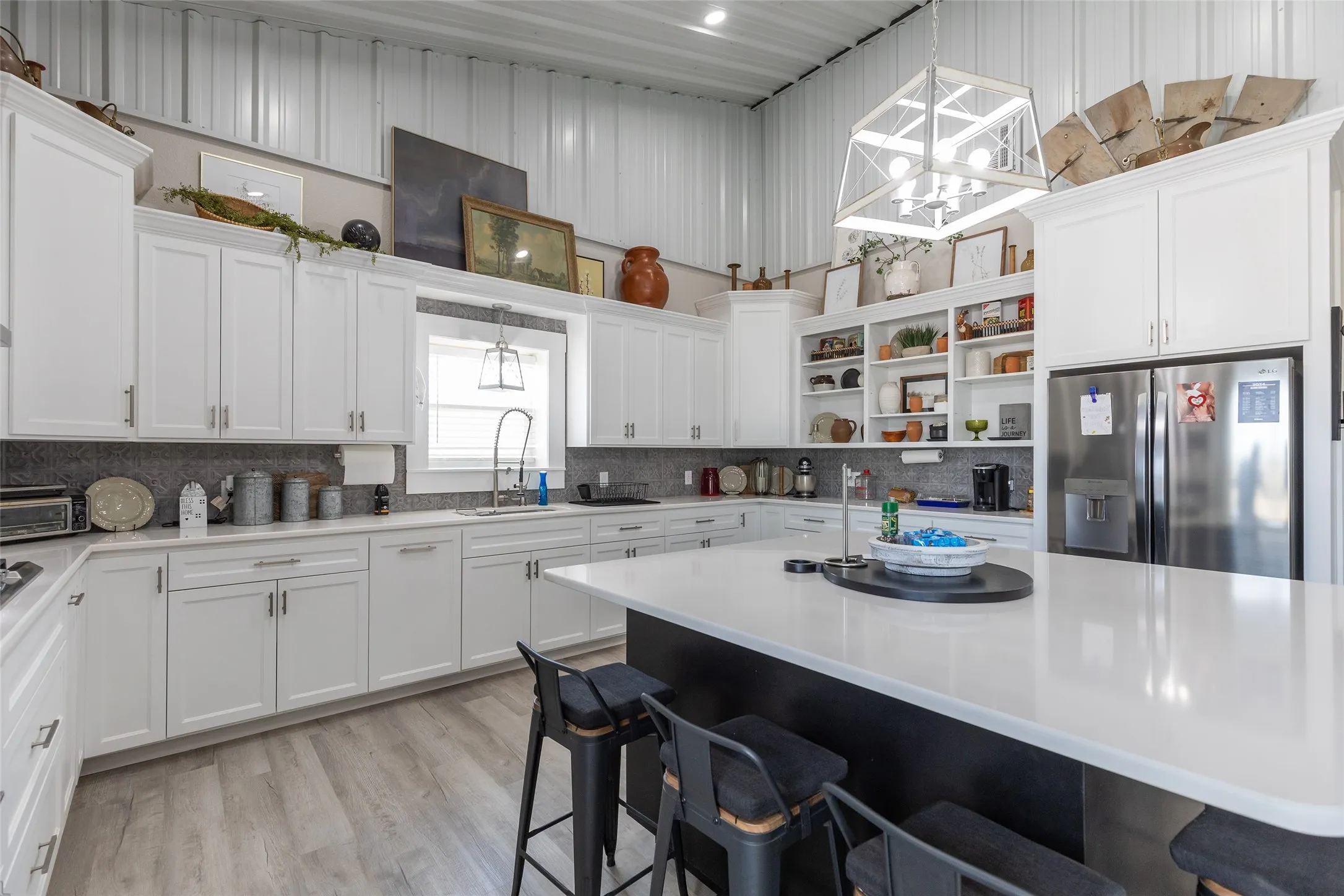 High ceilings with exposed metal beams, extensive white cabinetry, large center island with seating, stainless steel refrigerator, and wood-finish flooring