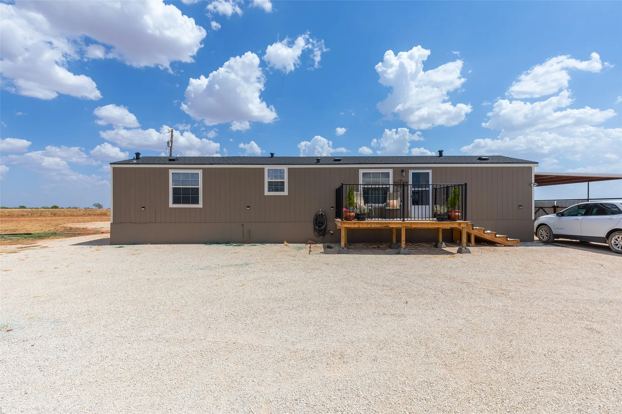 Exterior featuring tan siding, a wooden deck with black railings, a gravel driveway, white trim windows, and a covered carport structure