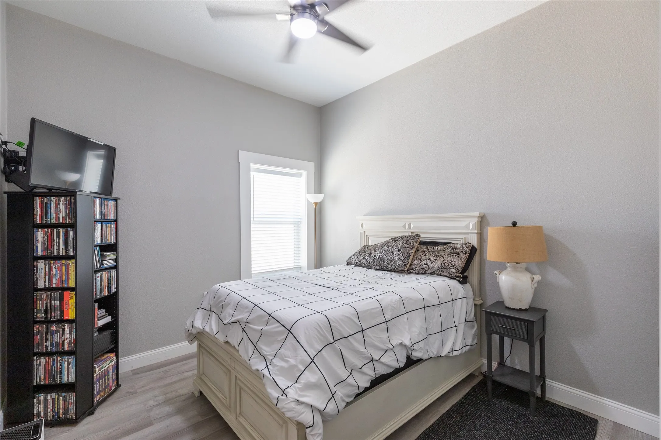 Light gray wall paint, wood-finish flooring, window with blinds, ceiling fan with integrated lighting, and white trim accents