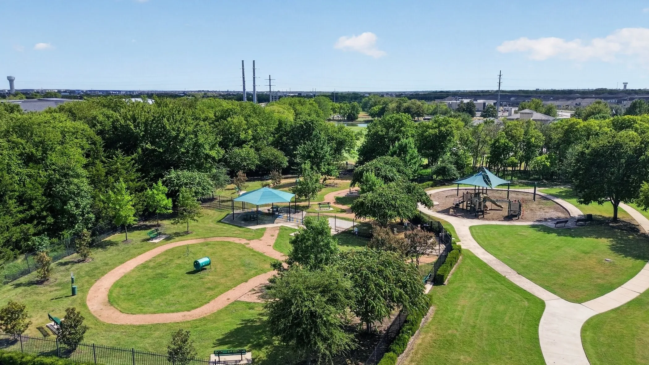 Expansive park setting featuring a playground, multiple covered pavilions, paved walking paths, and a dedicated pet exercise area