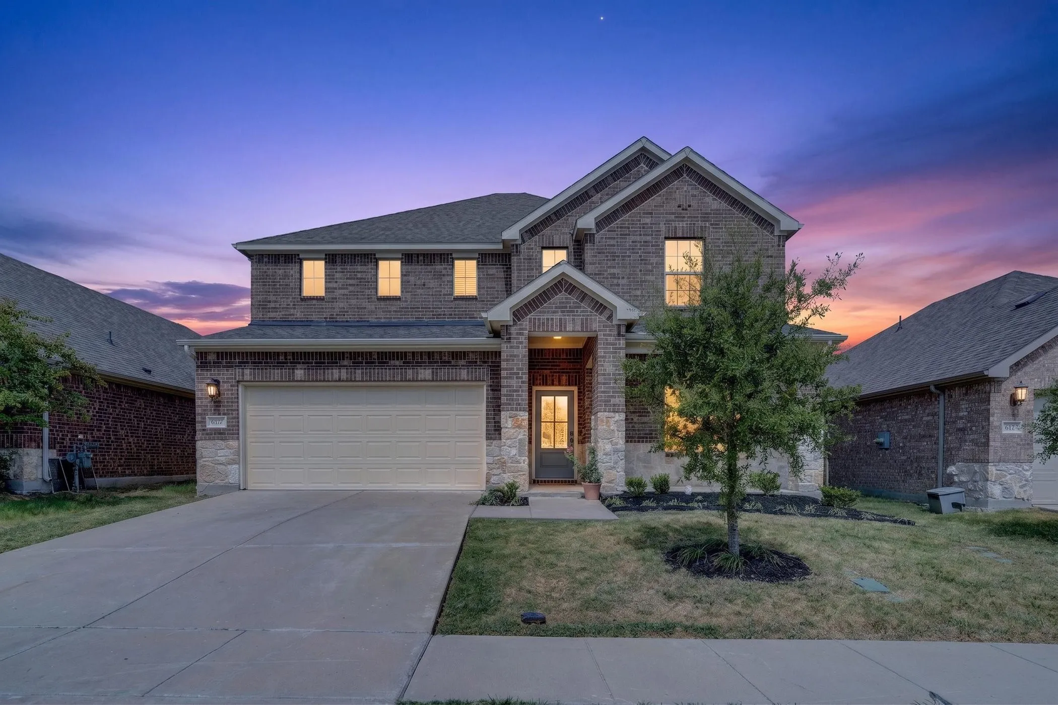 Two-story brick residence with welcoming front elevation featuring a two-car garage, illuminated multi-pane windows, and a mature landscape with established trees and lawn