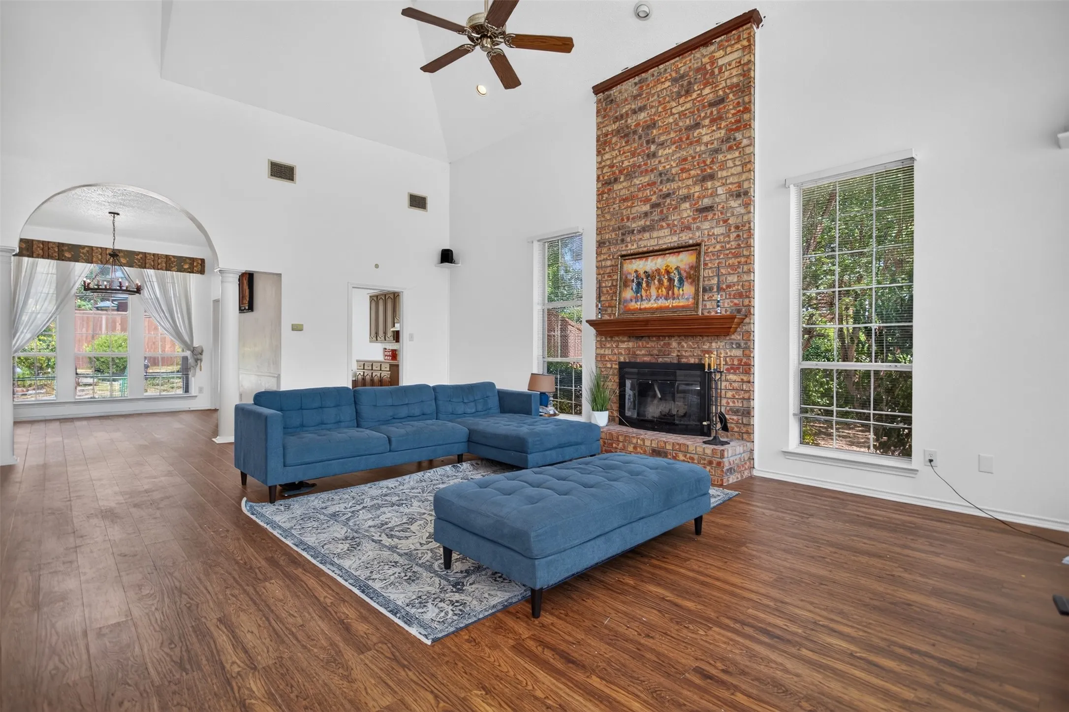 Vaulted ceiling with a ceiling fan, a brick-clad fireplace with a wood mantel, expansive windows, wood-finish flooring, and an arched entryway