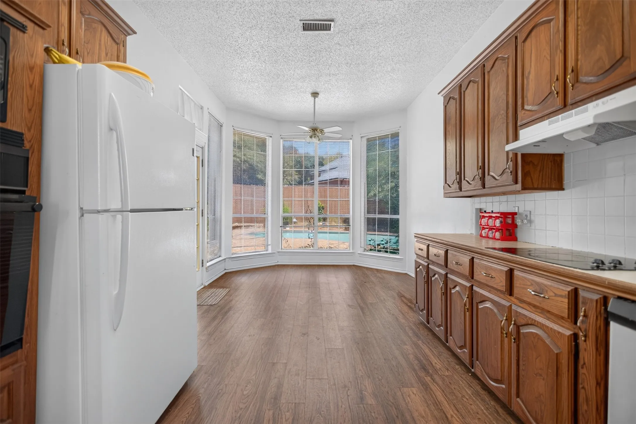 Wood-finish flooring throughout, bay windows overlooking the backyard, built-in cabinetry, tile backsplash, and a ceiling fan