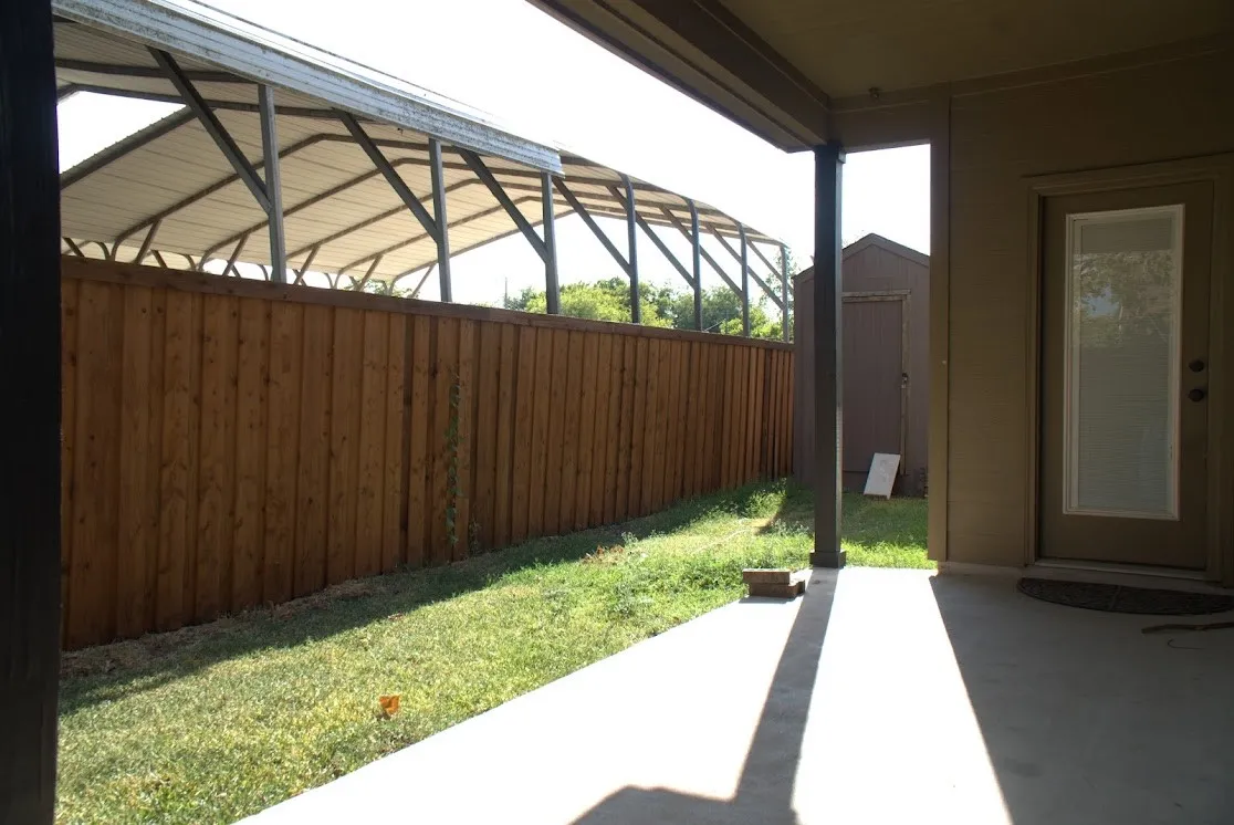 Covered patio with polished concrete flooring opening to a private backyard featuring a manicured lawn, wood-finish privacy fencing, and a steel-frame pergola structure providing shade and architectural interest