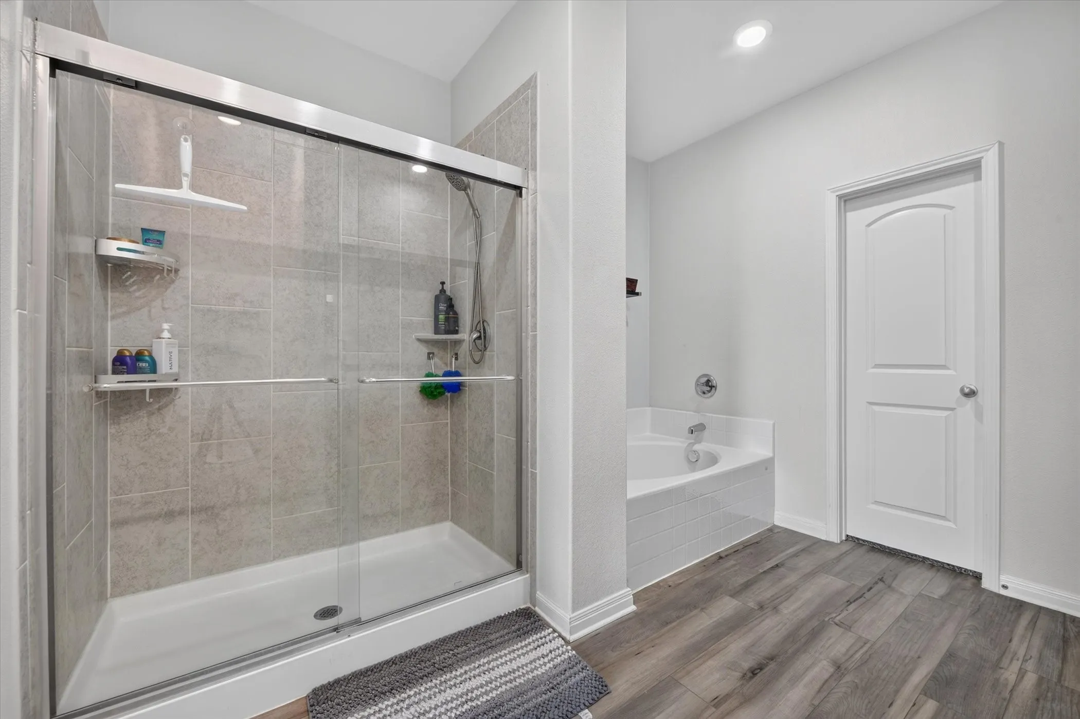 Walk-in shower with clear glass enclosure and tile surround, corner soaking tub, and wood-finish flooring